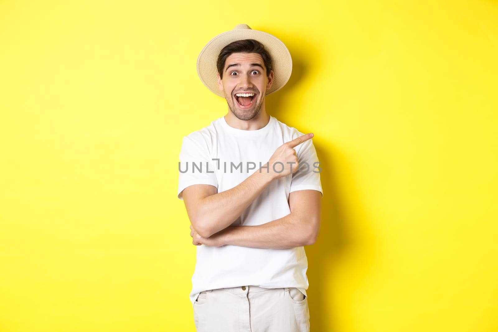 Happy male tourist in straw hat pointing finger right, showing promo offer on copy space, yellow background. Copy space