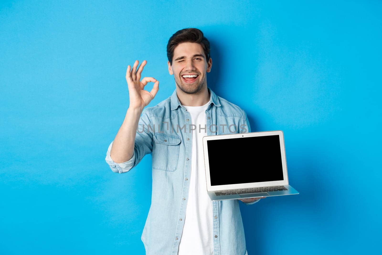 Young man showing laptop screen and okay sign, approve or like promo in internet, smiling satisfied, standing over blue background.