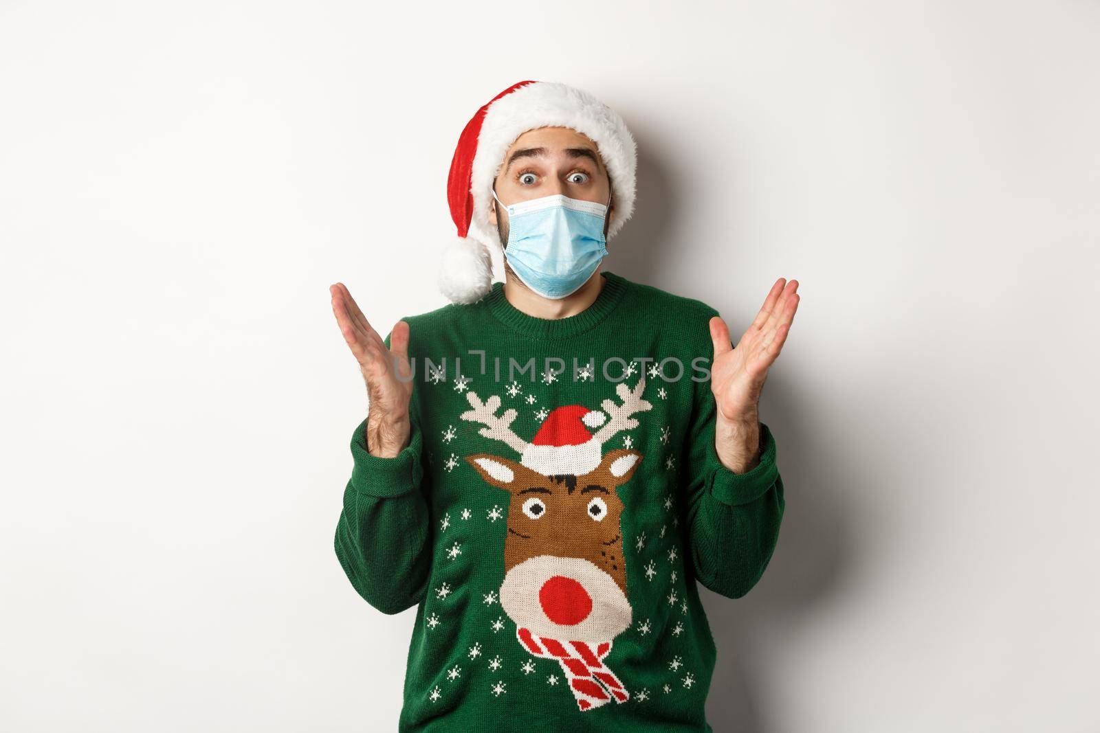 Christmas during pandemic, covid-19 concept. Surprised guy in medical mask, Santa hat and sweater celebrating New Year party, standing over white background.