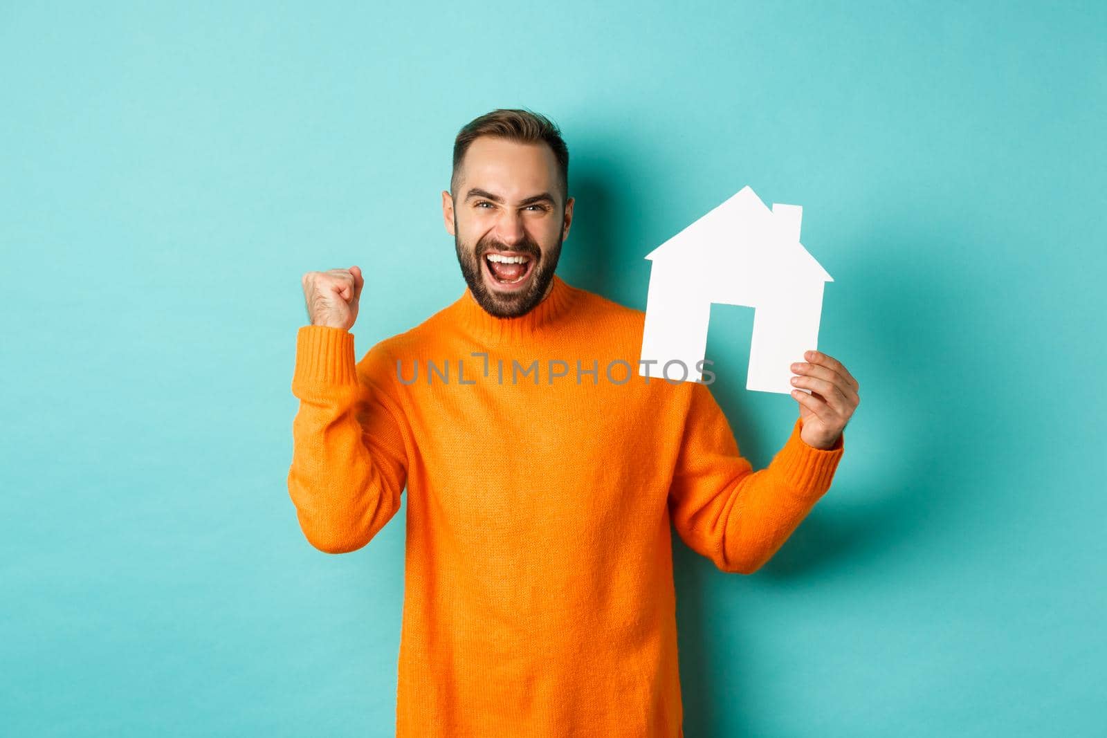 Real estate concept. Excited man saying yes, showing paper house maket and looking satisfied, standing in orange sweater over light blue background by Benzoix