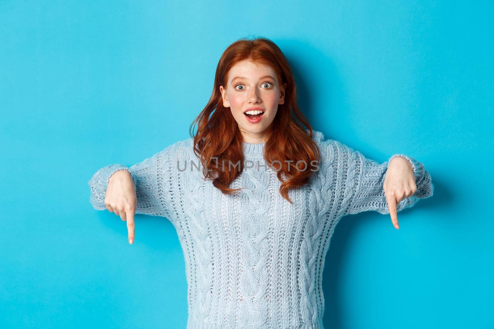 Winter holidays and people concept. Impressed and excited redhead girl, pointing fingers down and showing promo, staring camera amazed, standing over blue background.
