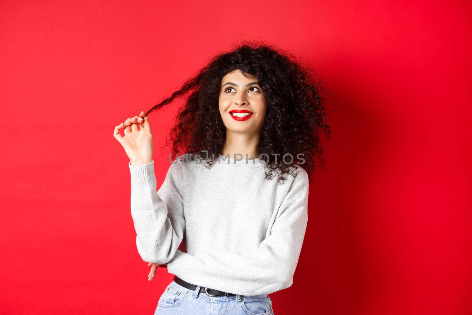 Beautiful female model playing with curly and looking happy up, thinking dreamy about something, standing against red background.