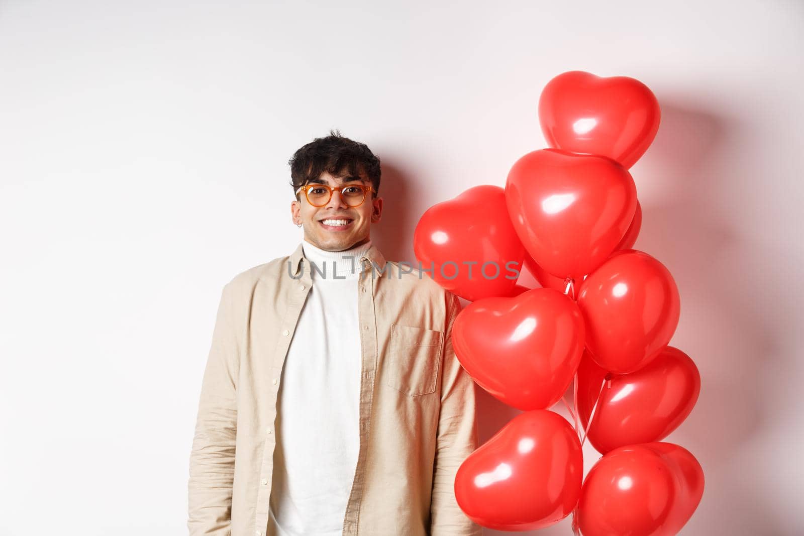 Valentines day. Excited young man smiling, looking hopeful, standing near big red hearts balloons, waiting for true love on lovers date, white background by Benzoix