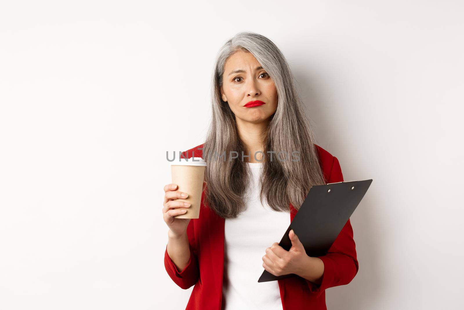 Tired and disappointed asian businesswoman with grey hair, drinking coffee in paper cup and looking gloomy at camera, standing over white background.