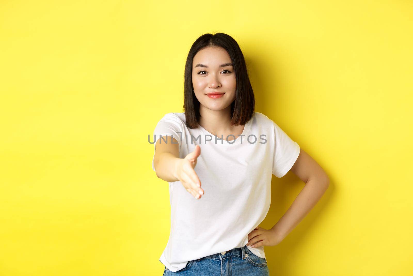 Confident asian woman in white t-shirt, stretch out hand for handshake and greeting gesture, saying hello, introduce herself, standing over yellow background.