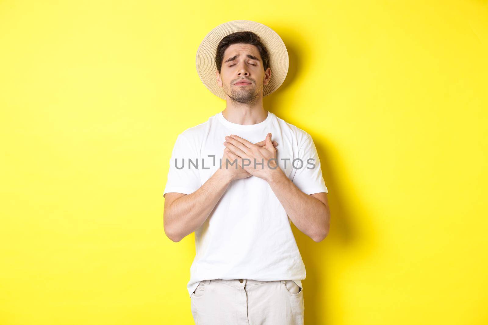 Concept of tourism and summer. Romantic man in straw hat looking nostalgic, close eyes and holding hands on heart, standing against yellow background.