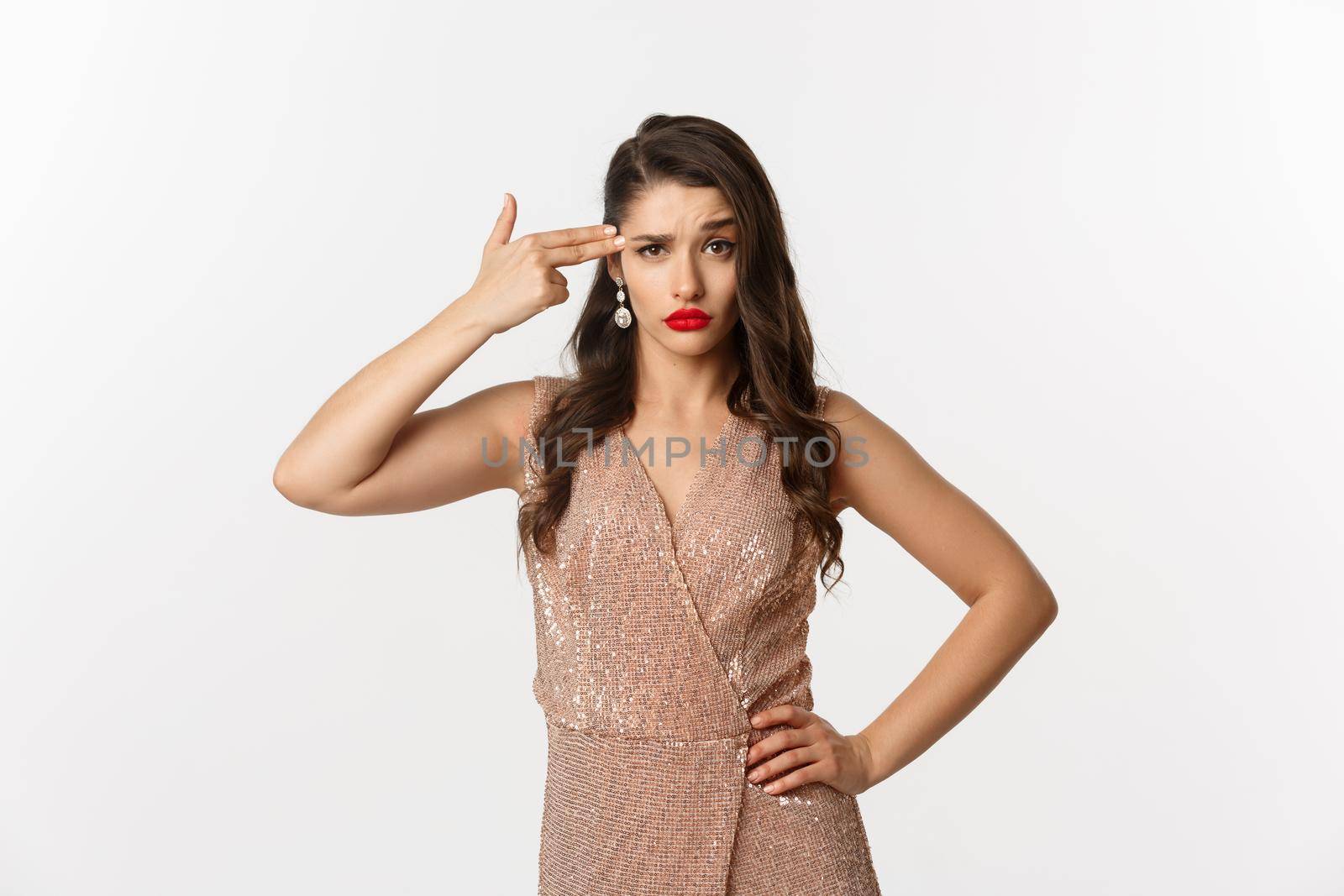 Celebration and party concept. Arrogant woman in elegant dress making finger gun at her head and looking bothered, standing annoyed over white background.