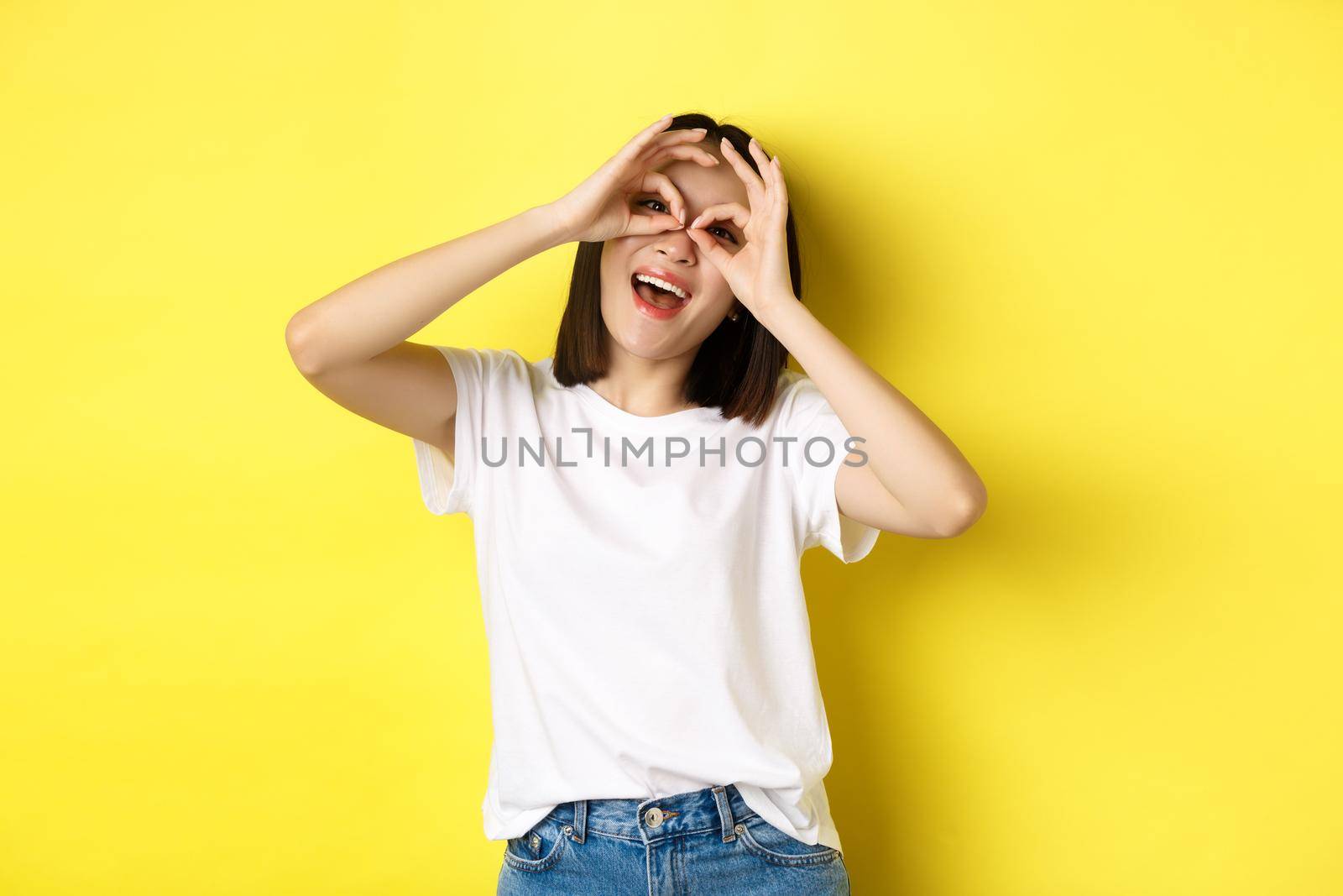 Funny asian girl looking through hand binoculars and smiling, stading over yellow background.