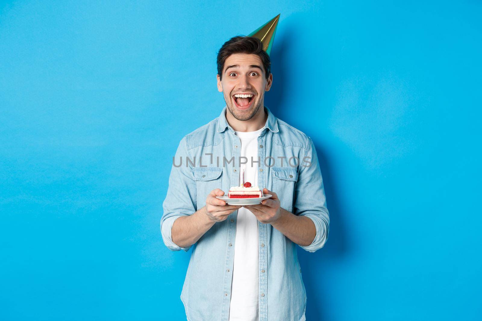 Cheerful young man celebrating birthday in party hat, holding b-day cake, standing against blue background.