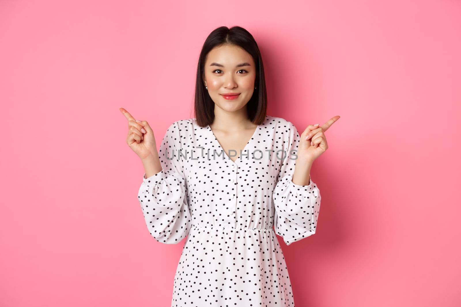 Beautiful asian woman making choice on shopping, pointing fingers sideways and showing variants, smiling at camera, standing over pink background.