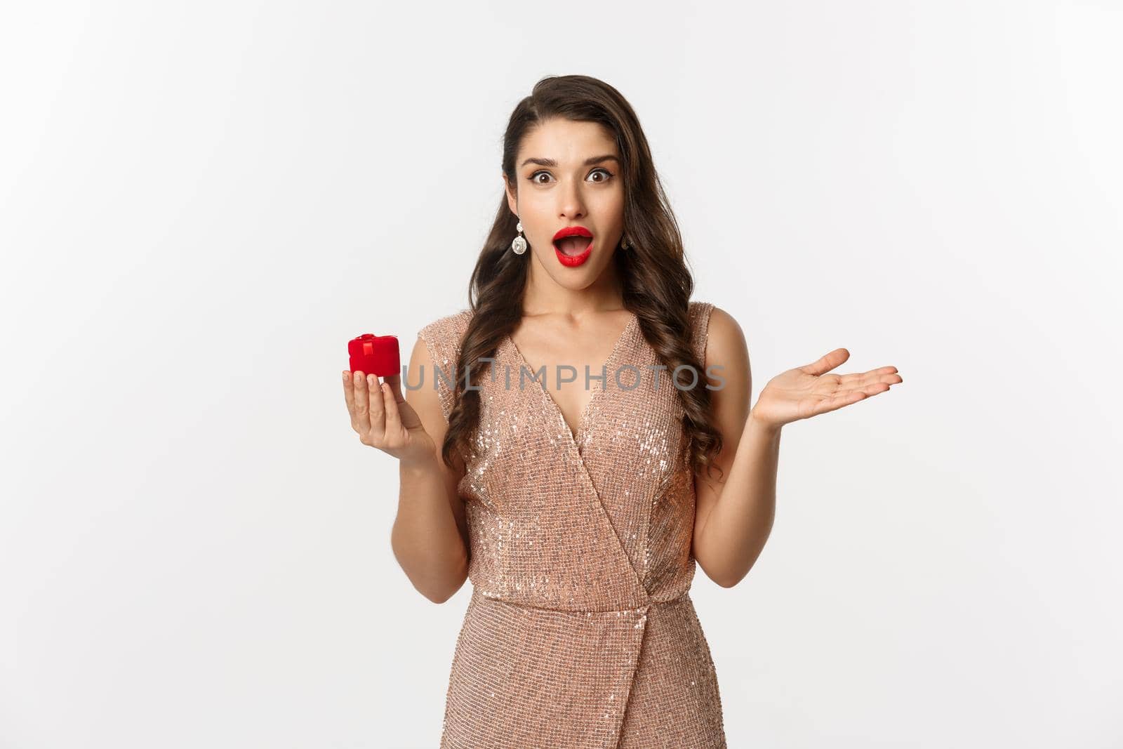 Image of amazed girlfriend holding engagement ring box, looking surprised, receive marriage proposal, wearing elegant dress, white background.