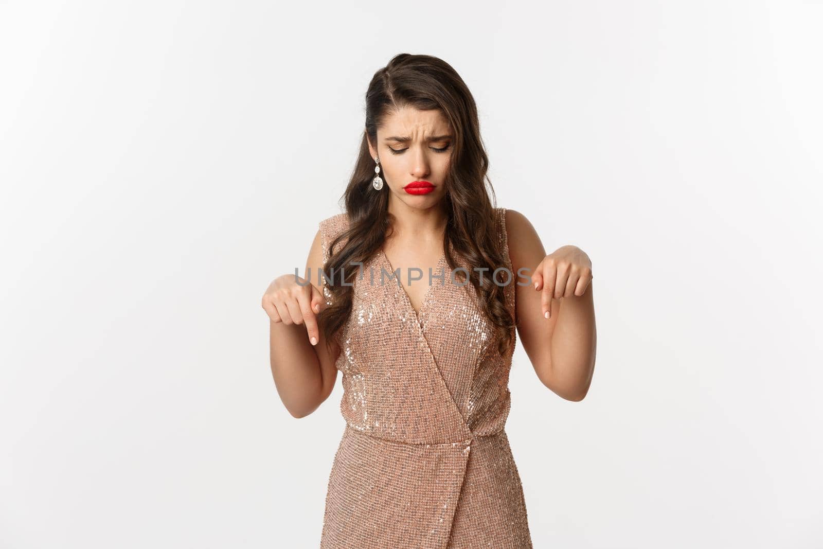 Sad beautiful girl pointing fingers down and sulking, wearing dress for christmas party, looking disappointed, standing over white background.