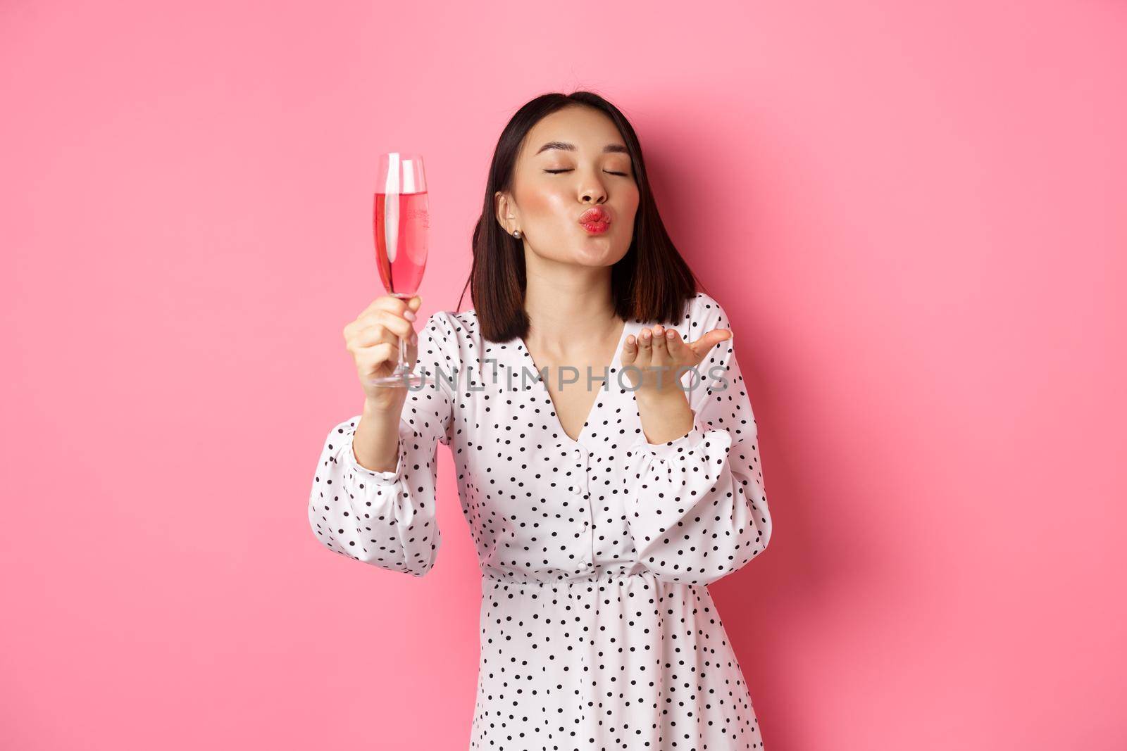 Romantic asian woman raising glass of champagne and sending air kiss at camera, celebrating and having fun, standing over pink background by Benzoix