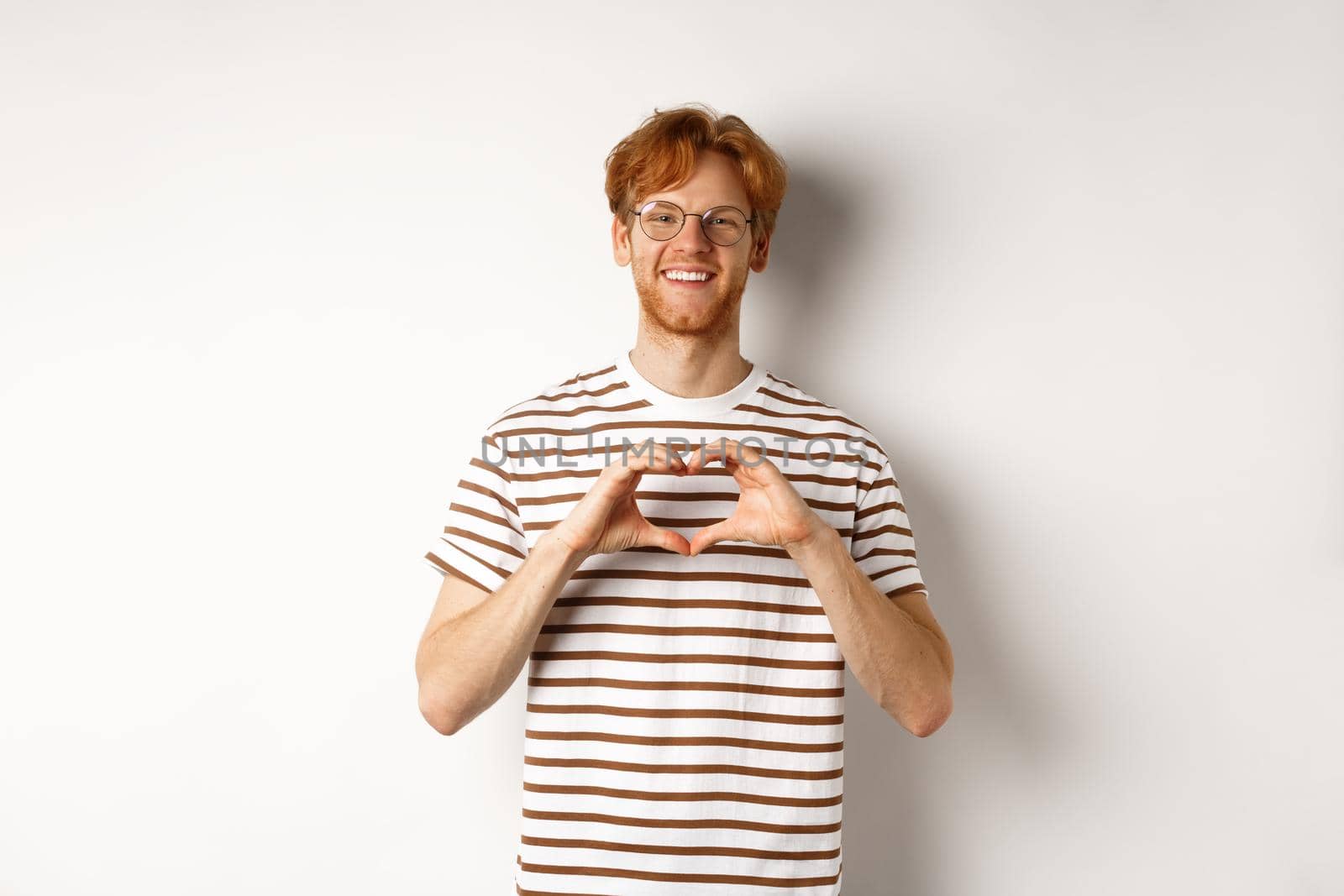 Valentines day. Happy boyfriend with red hair, smiling and showing heart gesture, I love you, standing over white background.
