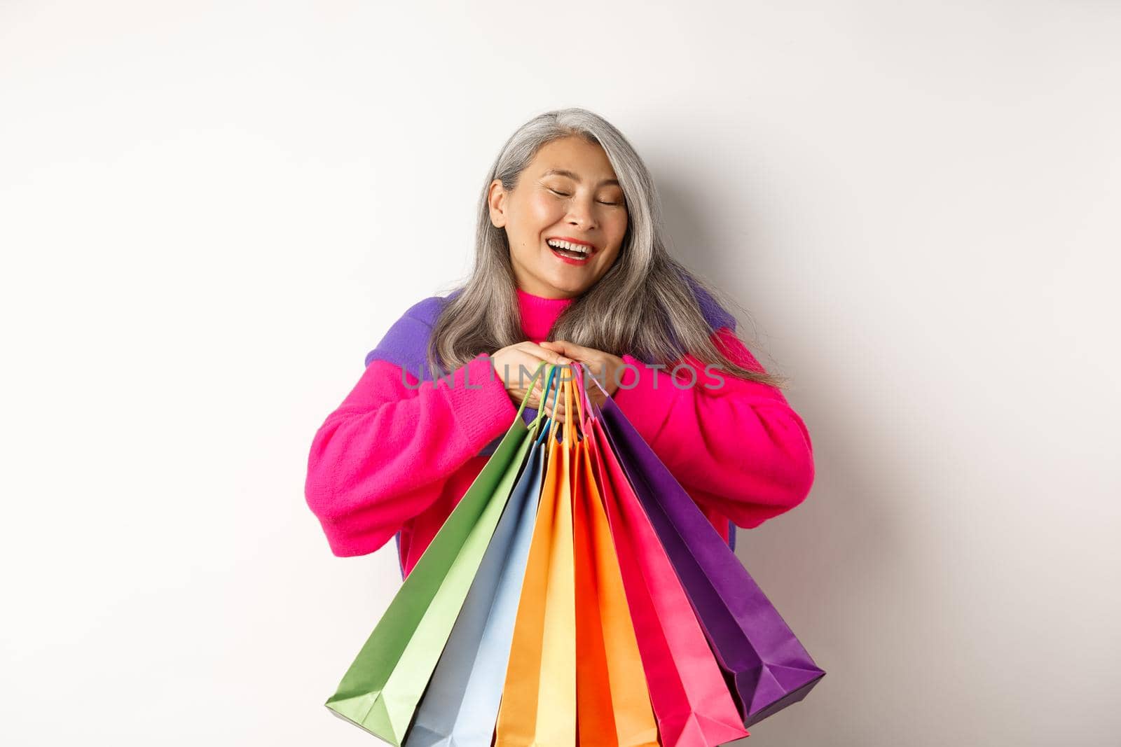 Fashionable senior asian woman shopaholic, hugging shopping bags and smiling joyful, buying with discounts, standing over white background by Benzoix