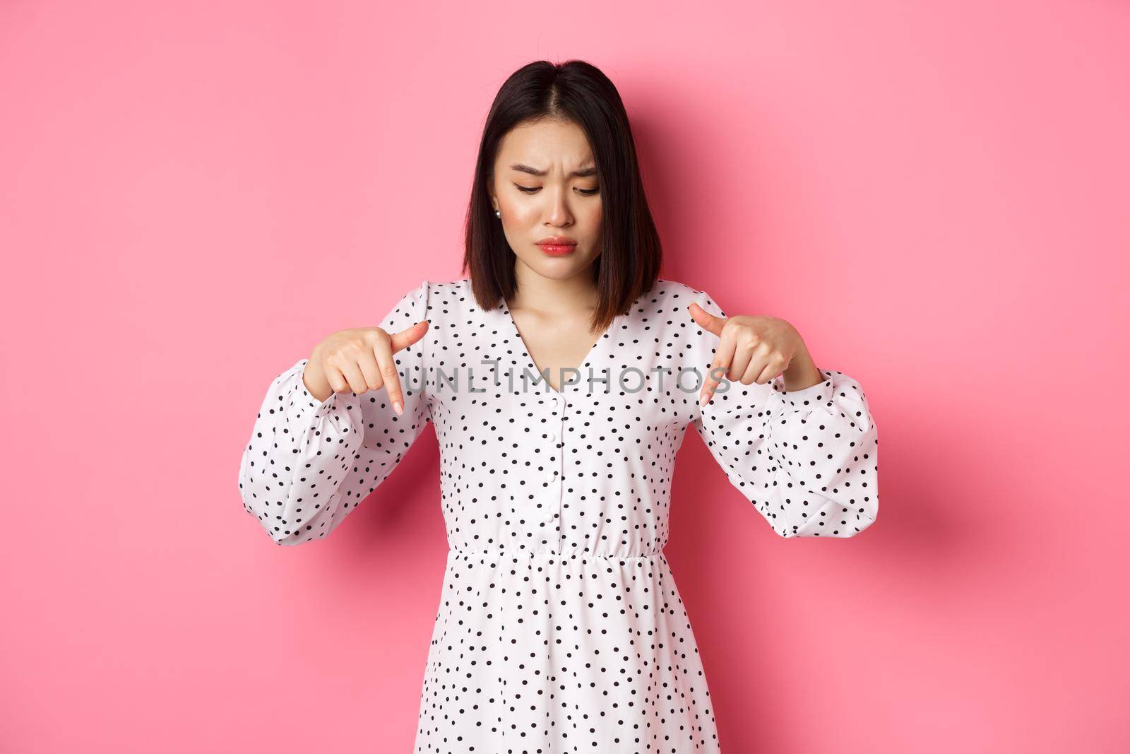 Concerned asian woman peeking down and pointing hand at disturbing banner, frowning upset, standing over pink background.