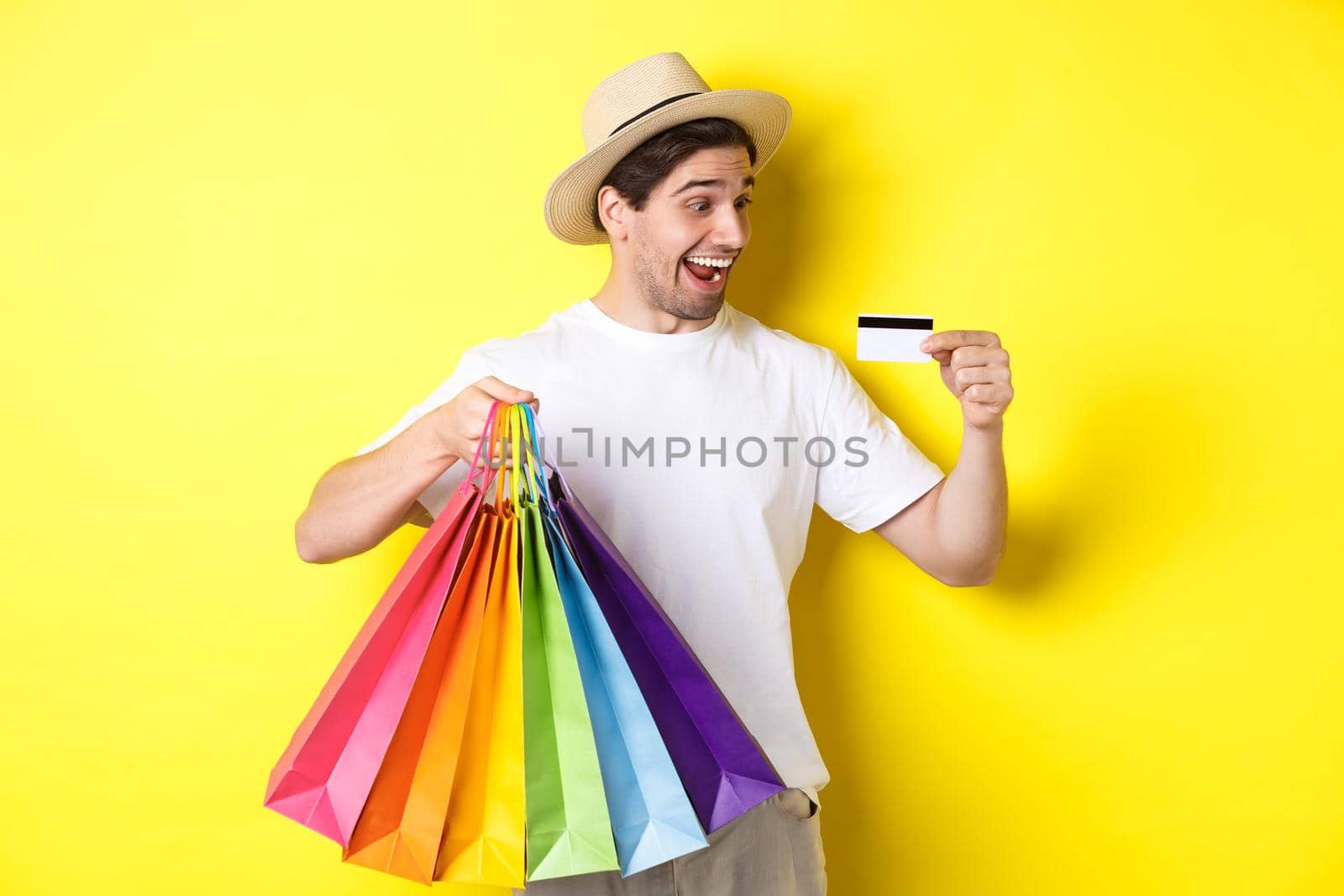 Excited man going shopping on vacation, looking satisfied at credit card, holding paper bags, standing over yellow background.