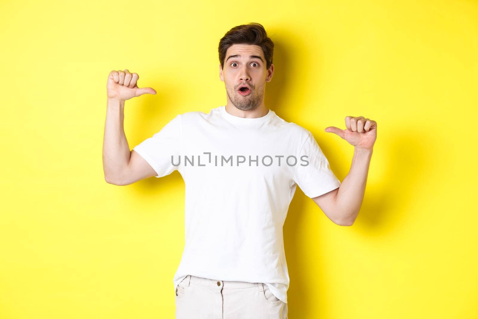 Surprised handsome guy pointing at himself, looking amazed, standing over yellow background. Copy space