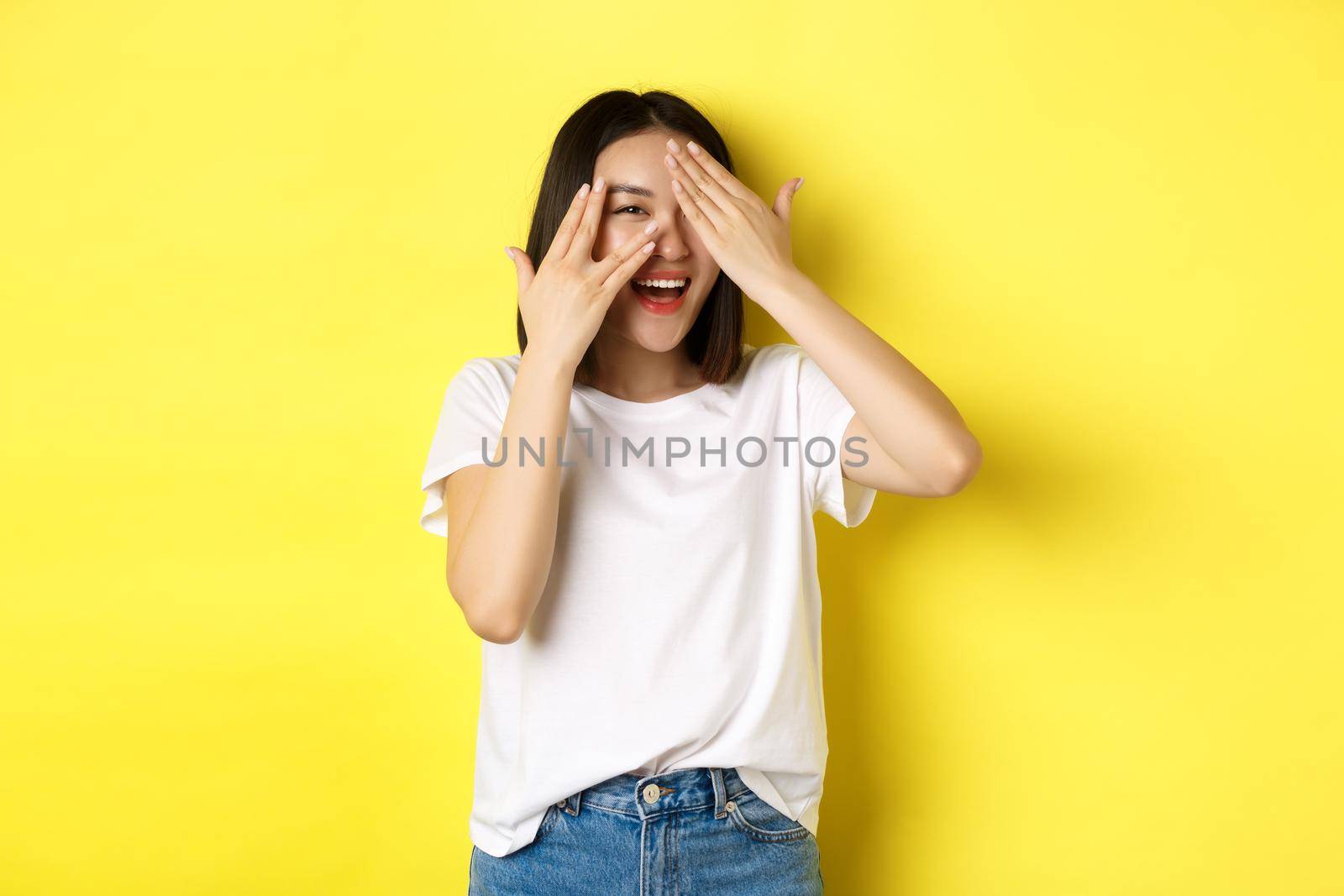 Intrigued asian girl waiting for surprise, peeking through fingers on eyes, smiling happy, standing over yellow background.