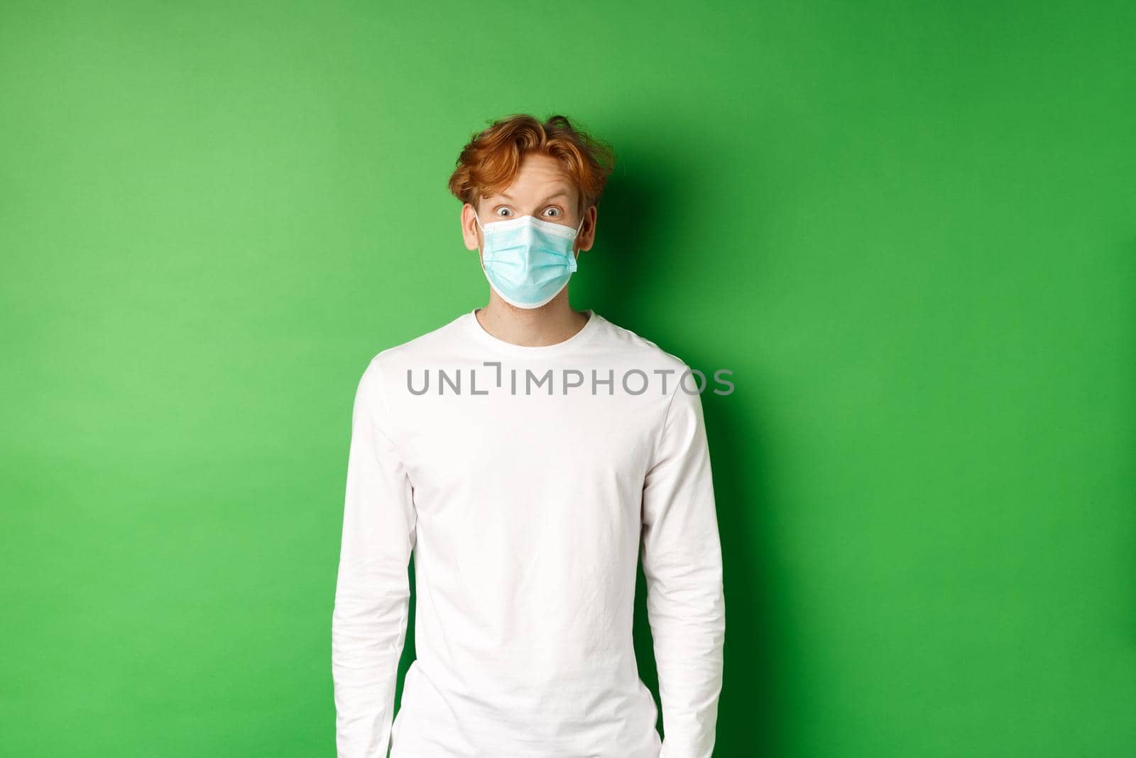 Covid-19, social distancing and lifestyle concept. Young redhead man wearing face mask during coronavirus pandemic, looking surprised at camera, green background.