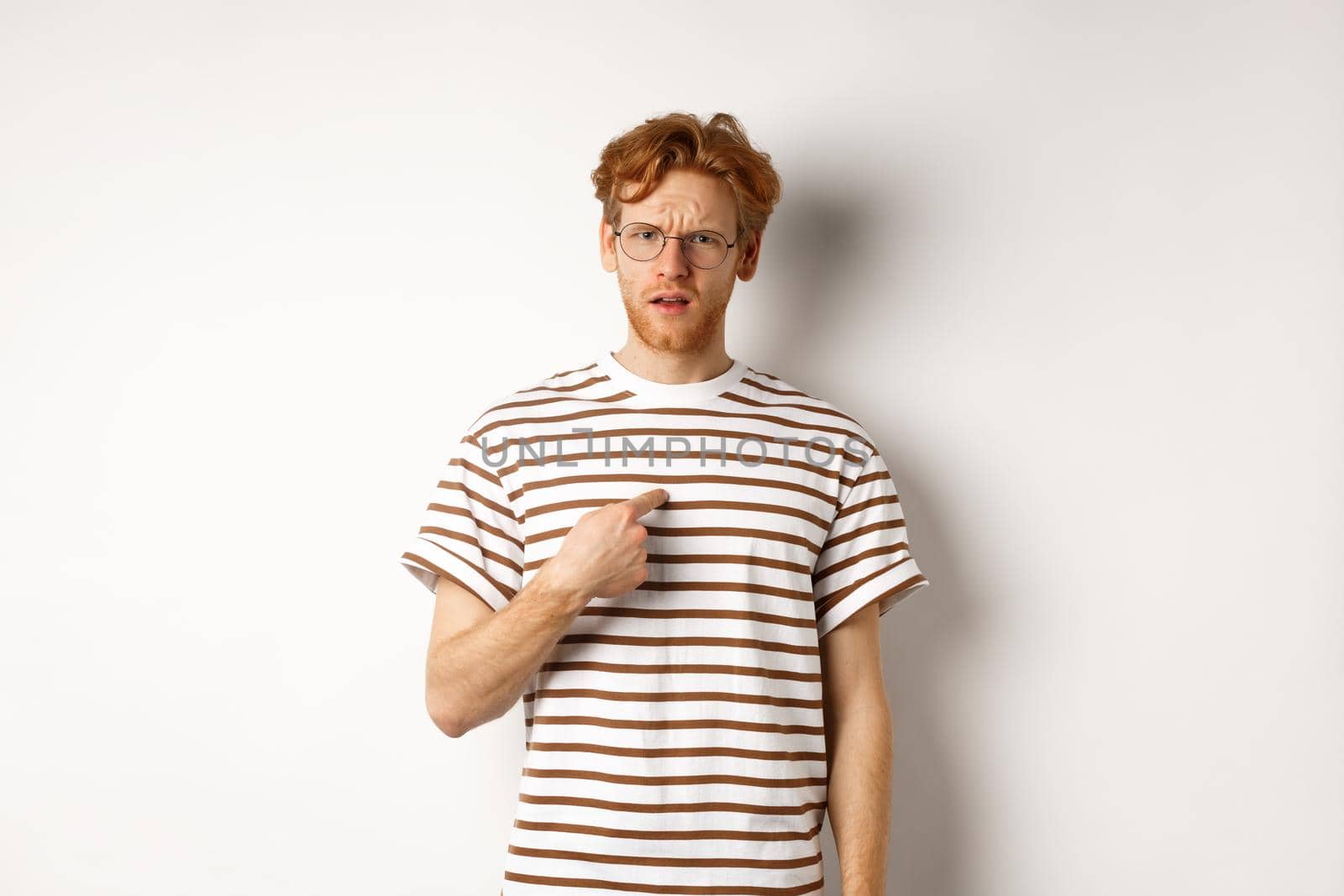 Confused redhead man in glasses pointing at himself, looking questioned at camera, white background.