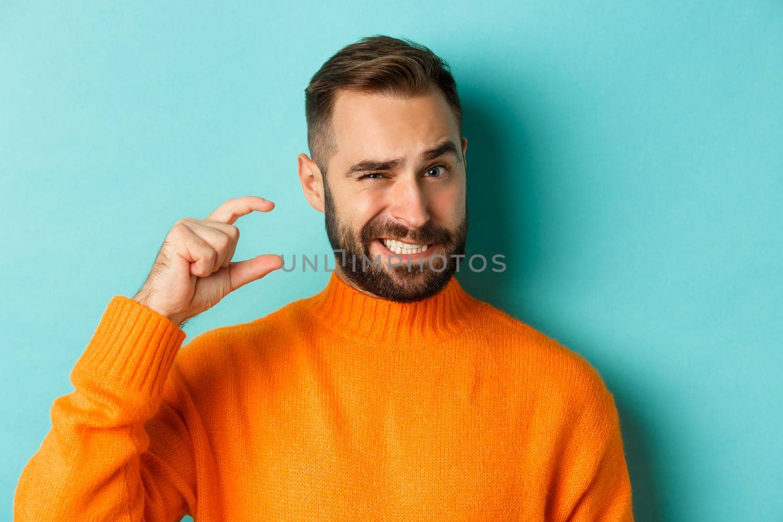 Close-up of awkward man showing small or little with fingers, standing displeased over light blue background.