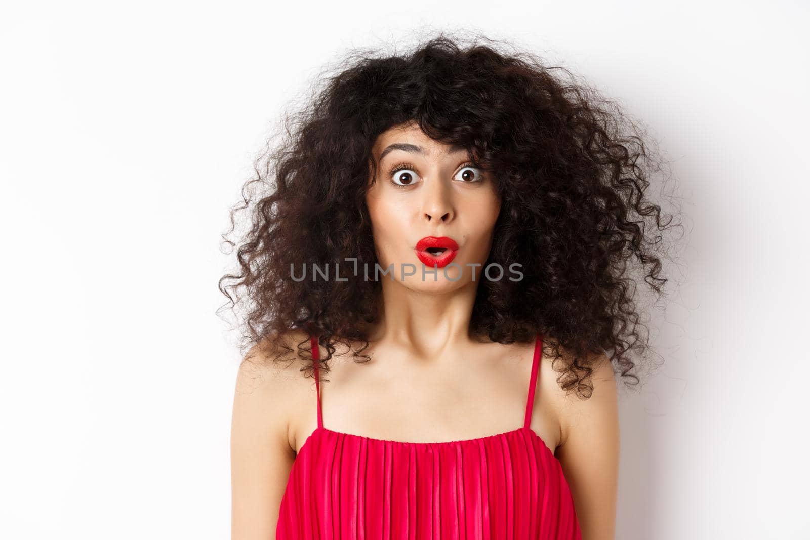 Close-up of surprised caucasian woman with red lips and curly hairstyle, saying wow and looking amazed, standing in red dress over white background.