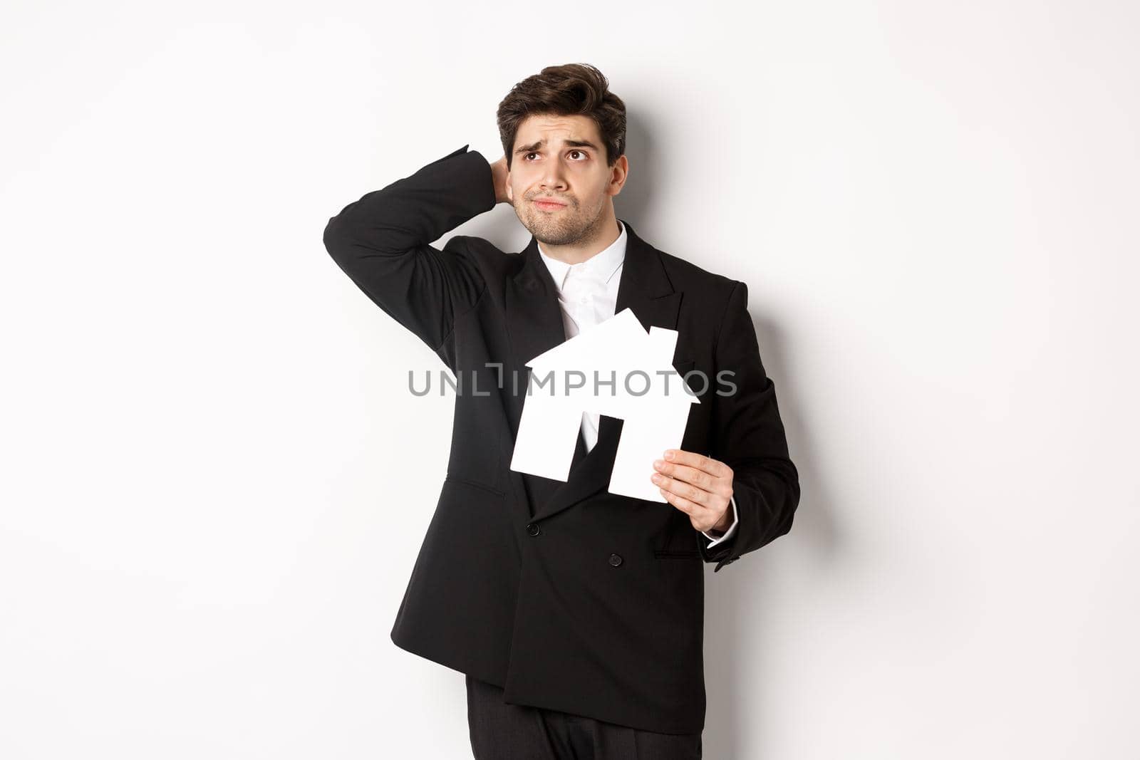 Image of indecisive businessman thinking, holding house maket and looking at upper left corner doubtful, standing against white background by Benzoix