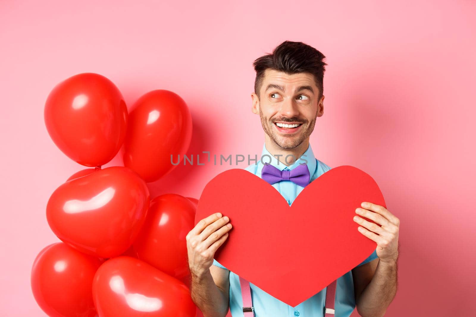 Valentines day concept. Romantic guy smiling and looking left, dreaming of date with lover, showing red big heart cutout, pink background.