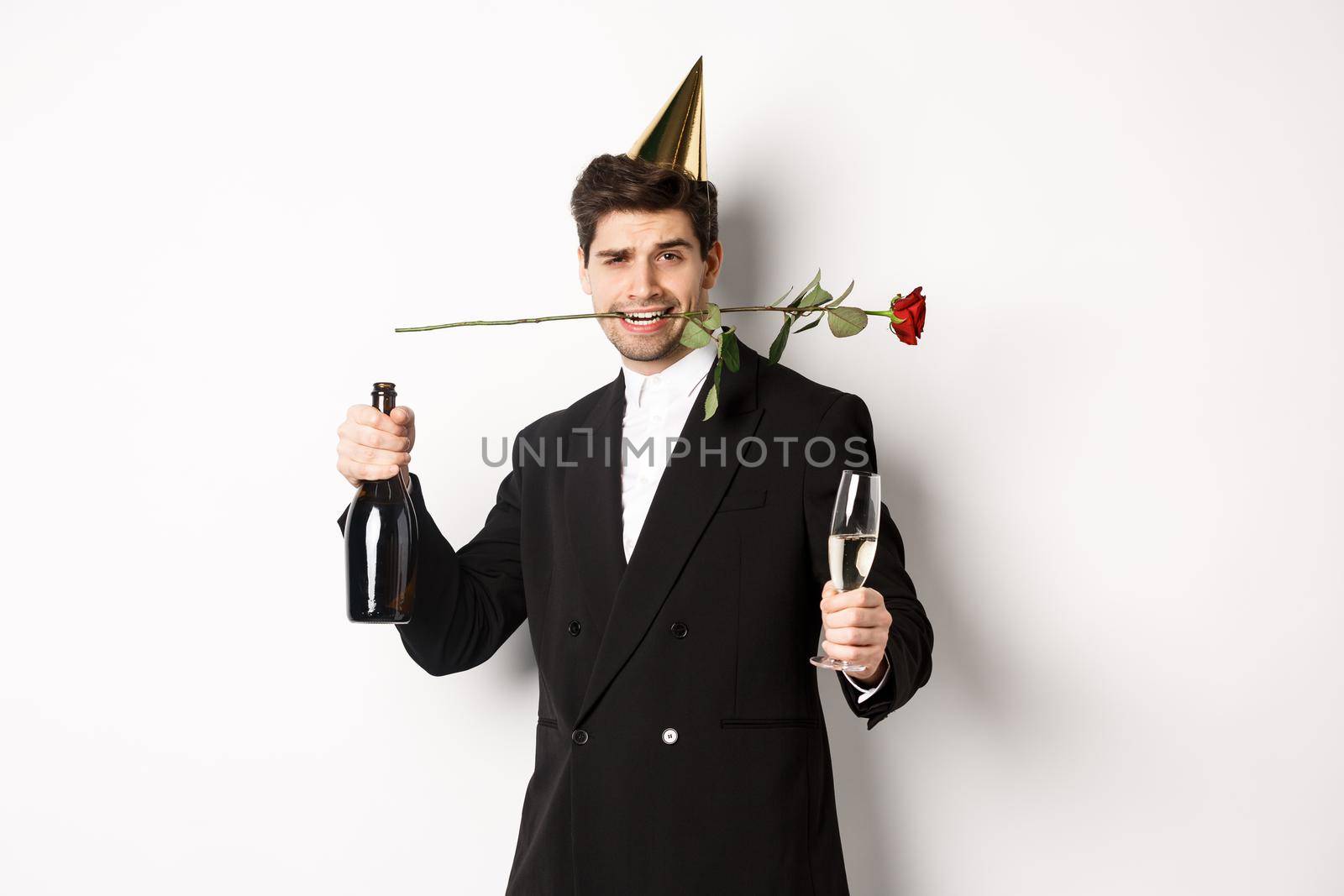 Romantic guy in trendy suit, celebrating and having a party, holding rose in teeth and champagne, standing over white background.