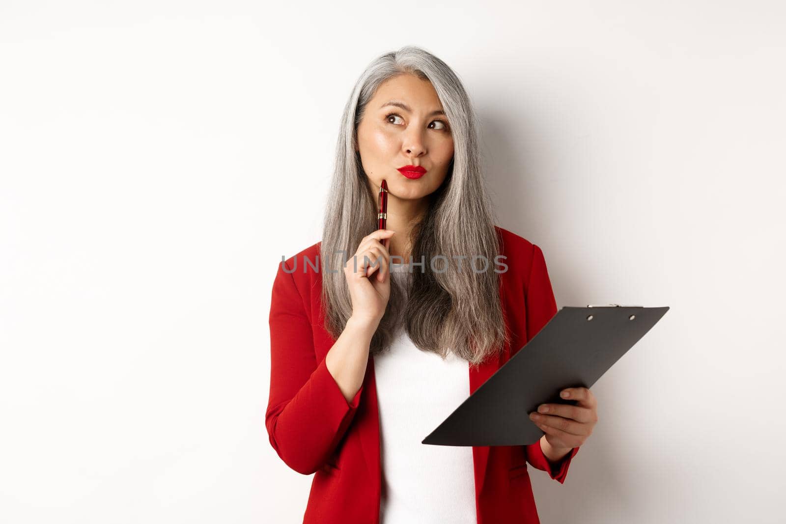 Stylish asian office lady in red blazer writing report on clipboard, looking thoughtful while working, standing over white background.