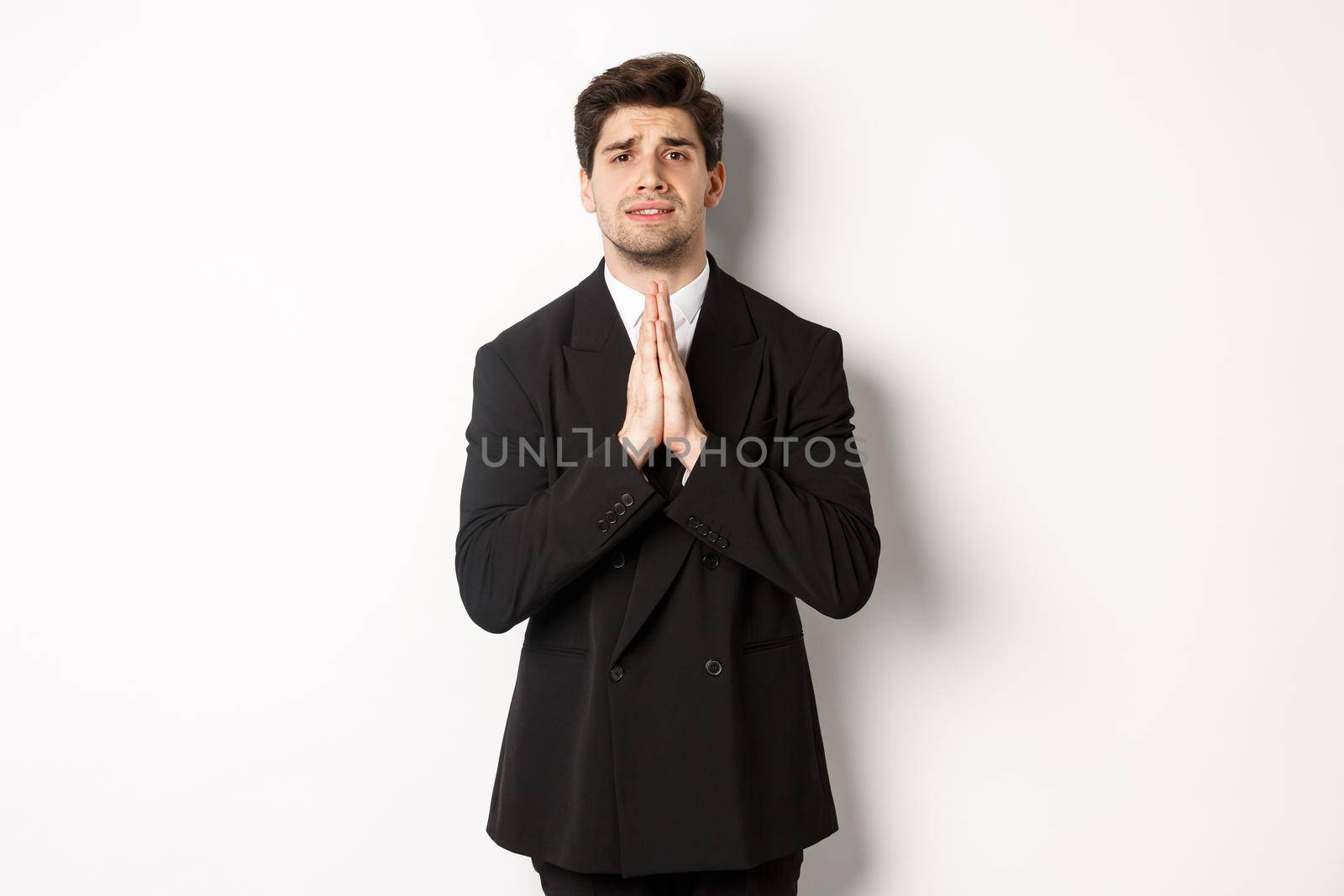 Image of handsome businessman in suit need help, begging you, holding hands in plead gesture and looking desperate, standing over white background.