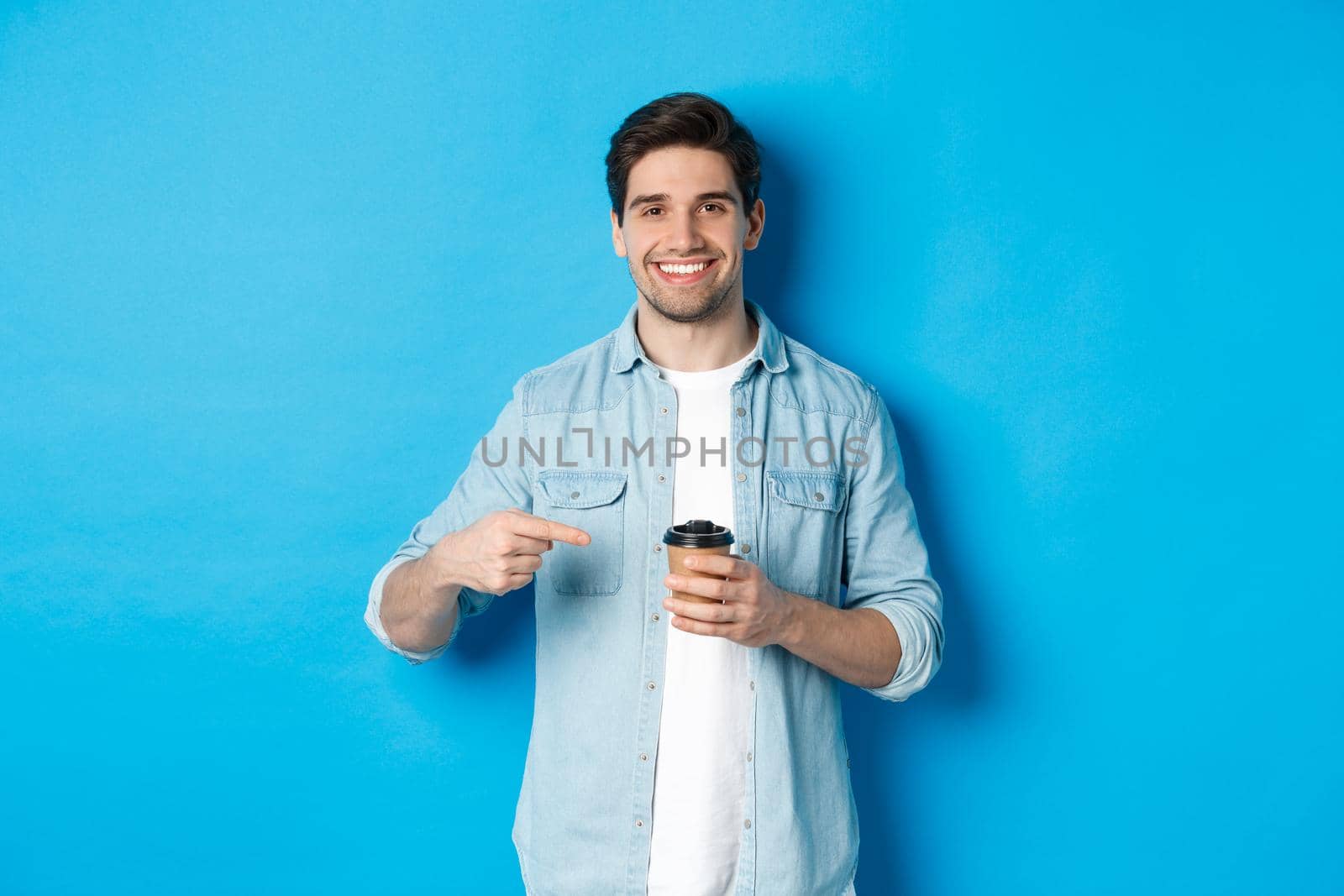 Smiling happy man pointing at paper cup with coffee, recommending cafe, standing over blue background.