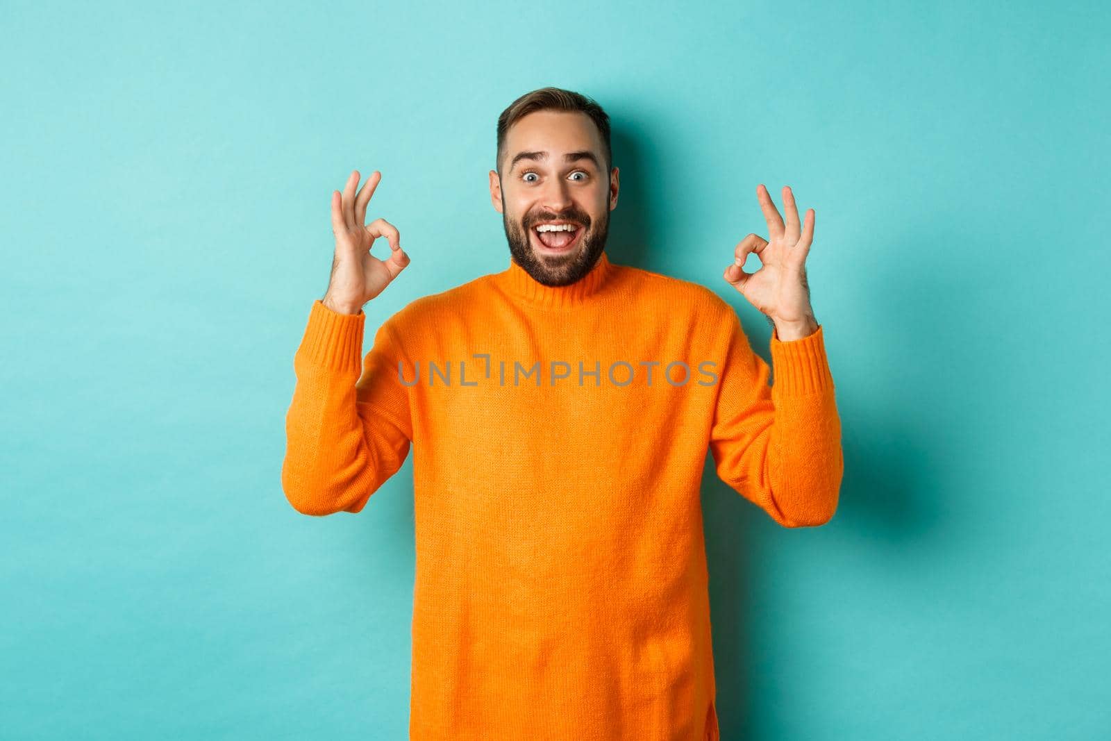 Impressed man praising something awesome, showing okay signs and looking amazed, standing in orange sweater at light blue background.