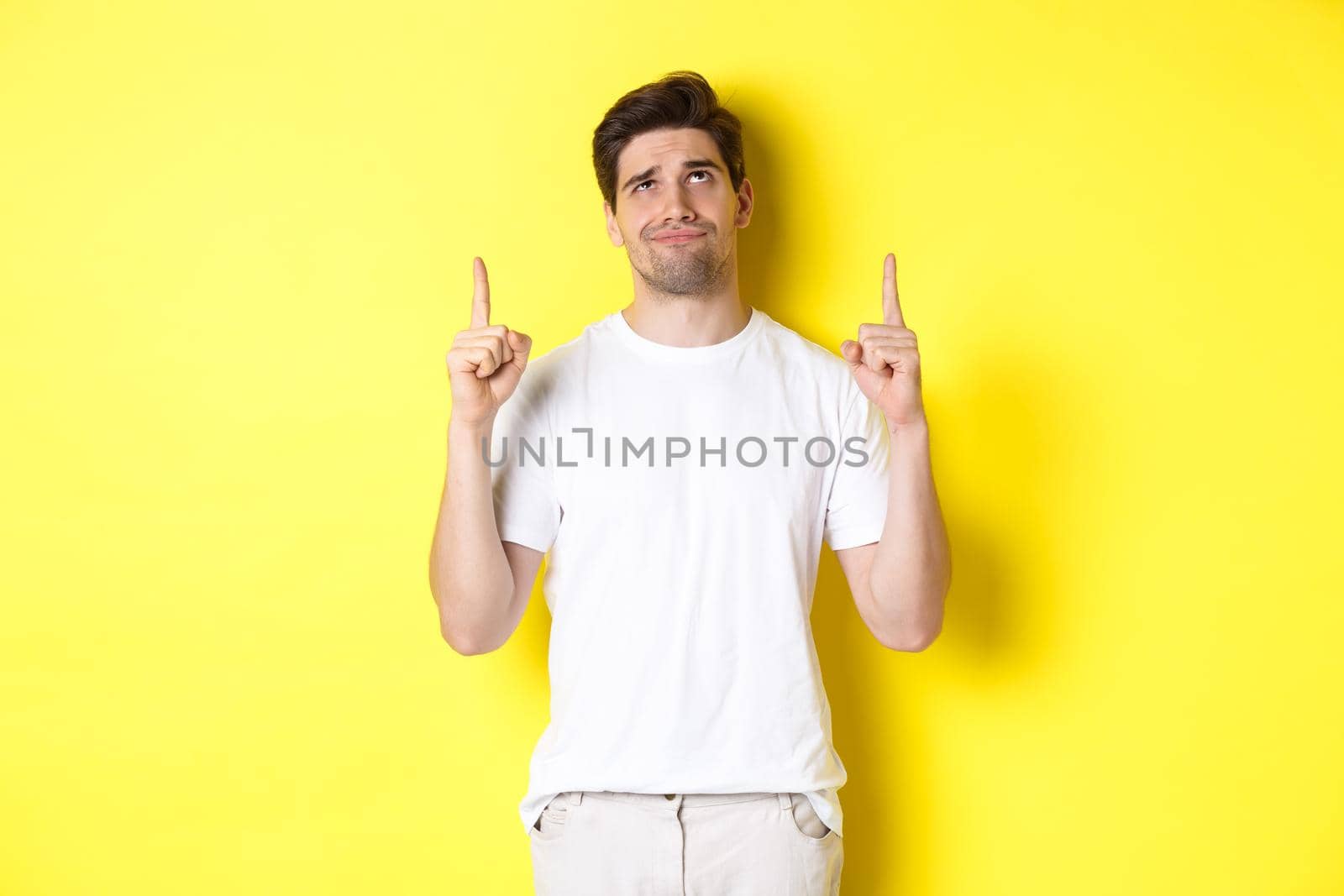 Skeptical young man pointing and looking up at something bad, judging offer, standing over yellow background by Benzoix