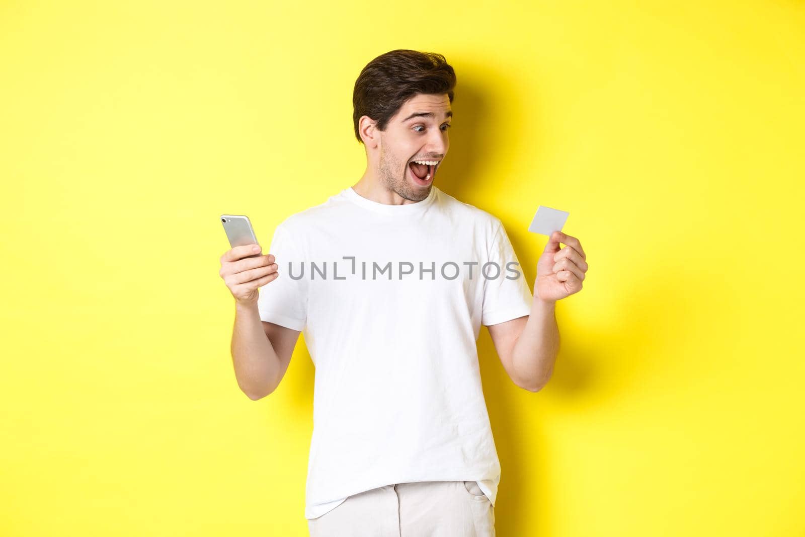Surprised guy holding smartphone and credit card, online shopping on black friday, standing over yellow background.