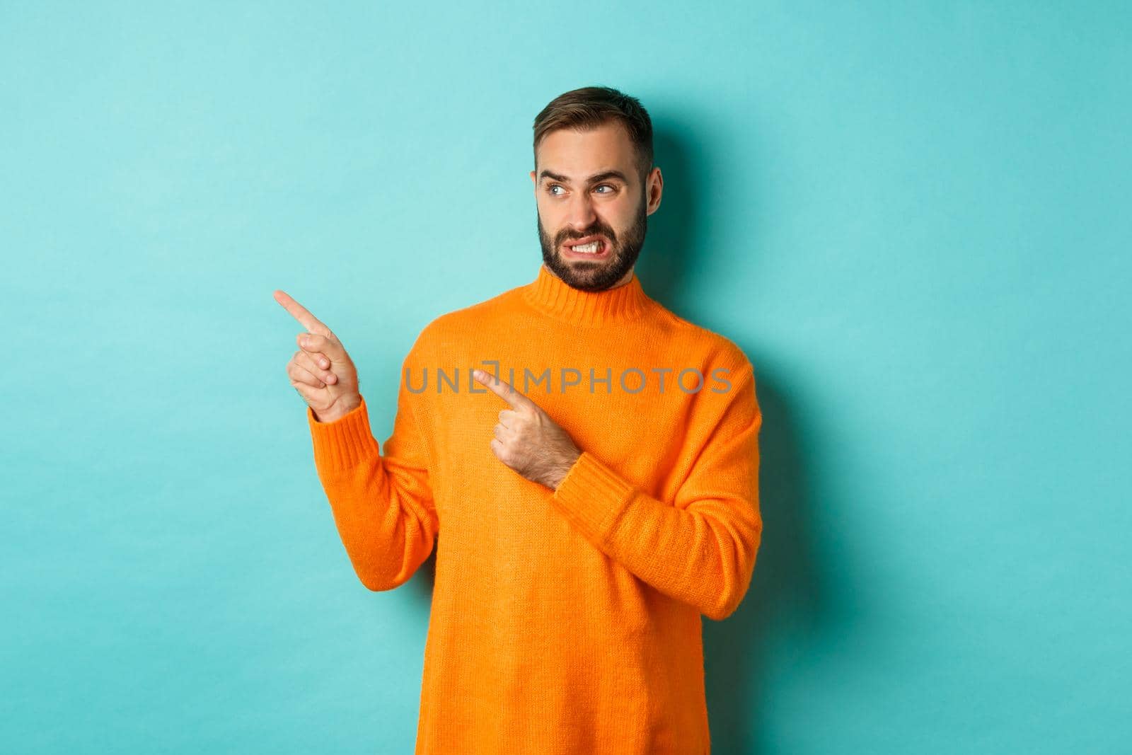 Displeased man cringe from something bad, pointing and looking at something disgusting or strange, left side copy space, standing against turquoise background by Benzoix