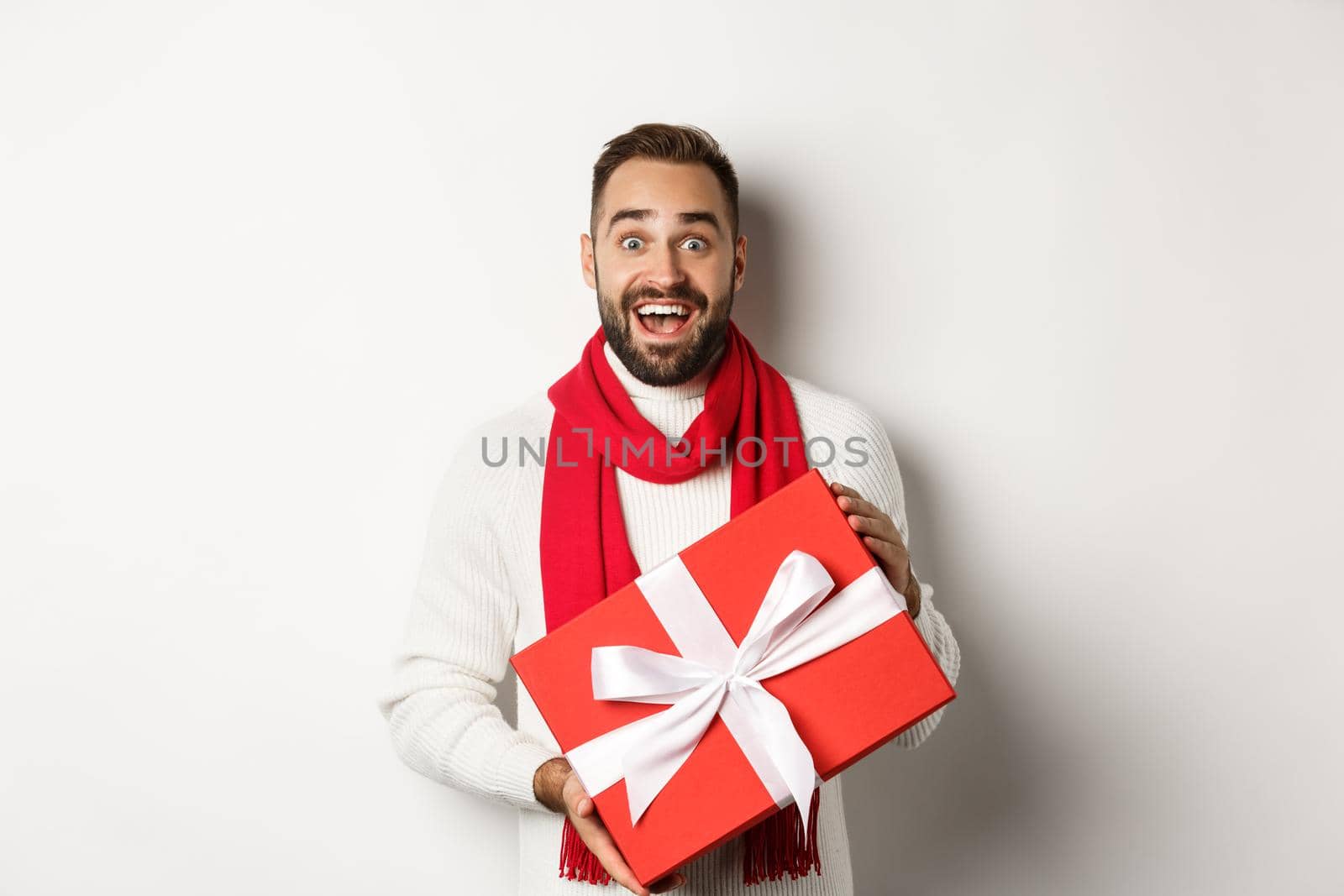 Concept of winter holidays. Surprised man receiving christmas gift and looking amazed, holding red wrapped box with present, standing amazed at white background.