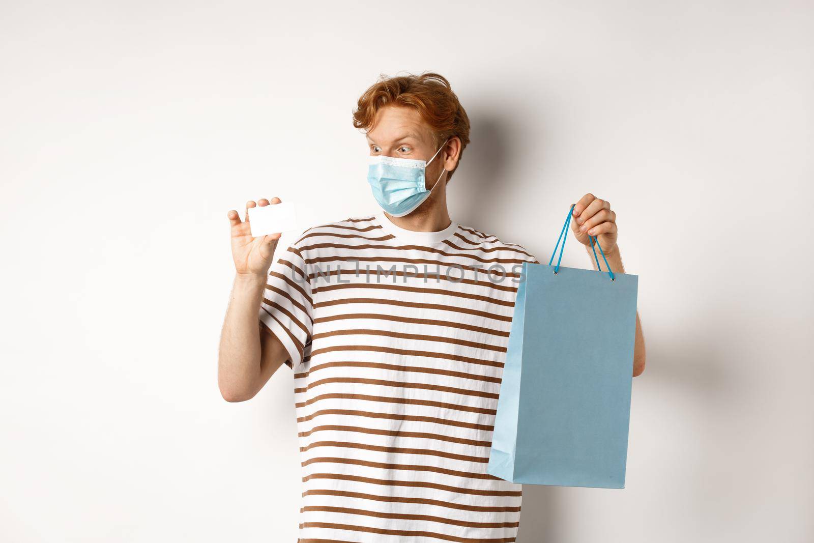 Concept of covid-19 and lifestyle. Cheerful young man with red hair, wear medical mask, showing shopping bag from store and plastic credit card.