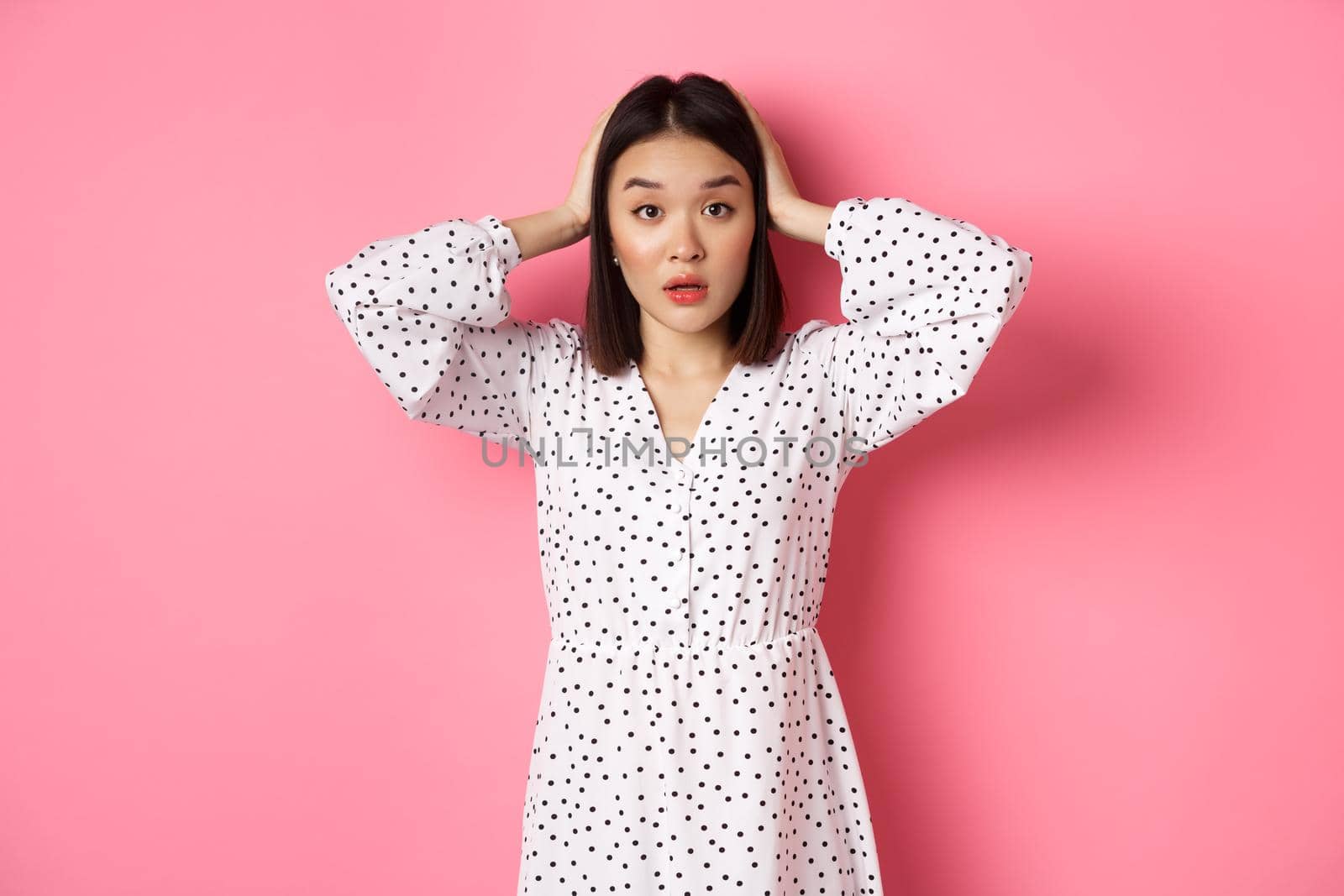 Shocked and troubled asian woman looking confused, holding hands on head, having problem, standing over pink background.