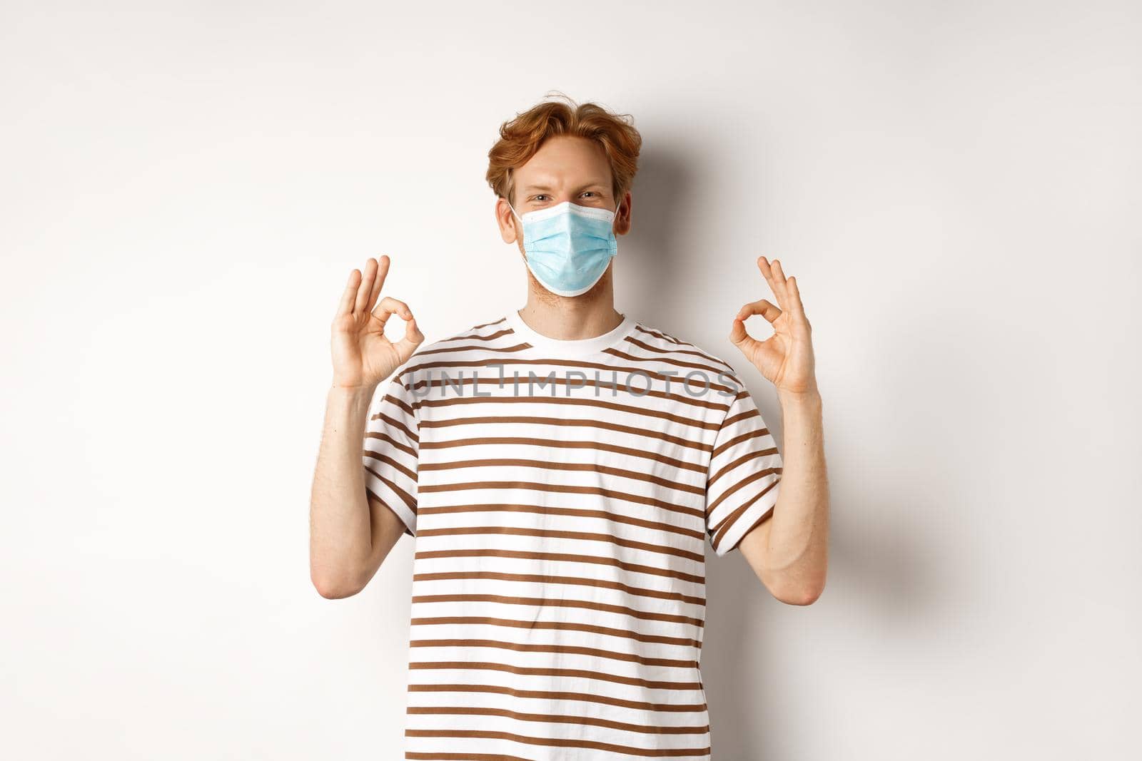 Covid-19, pandemic and social distancing concept. Young man with red hair wearing medical mask to prevent catching coronavirus, showing okay signs in approval, white background.