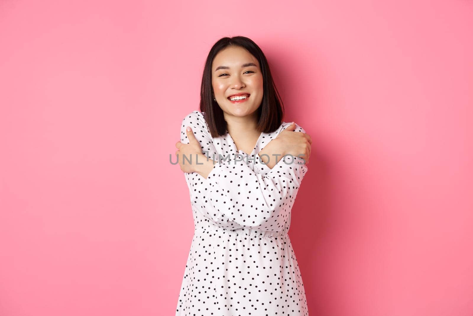 Beautiful asian girl embracing own body, hugging herself and smiling with happy face, looking at camera carefree, standing over pink background by Benzoix