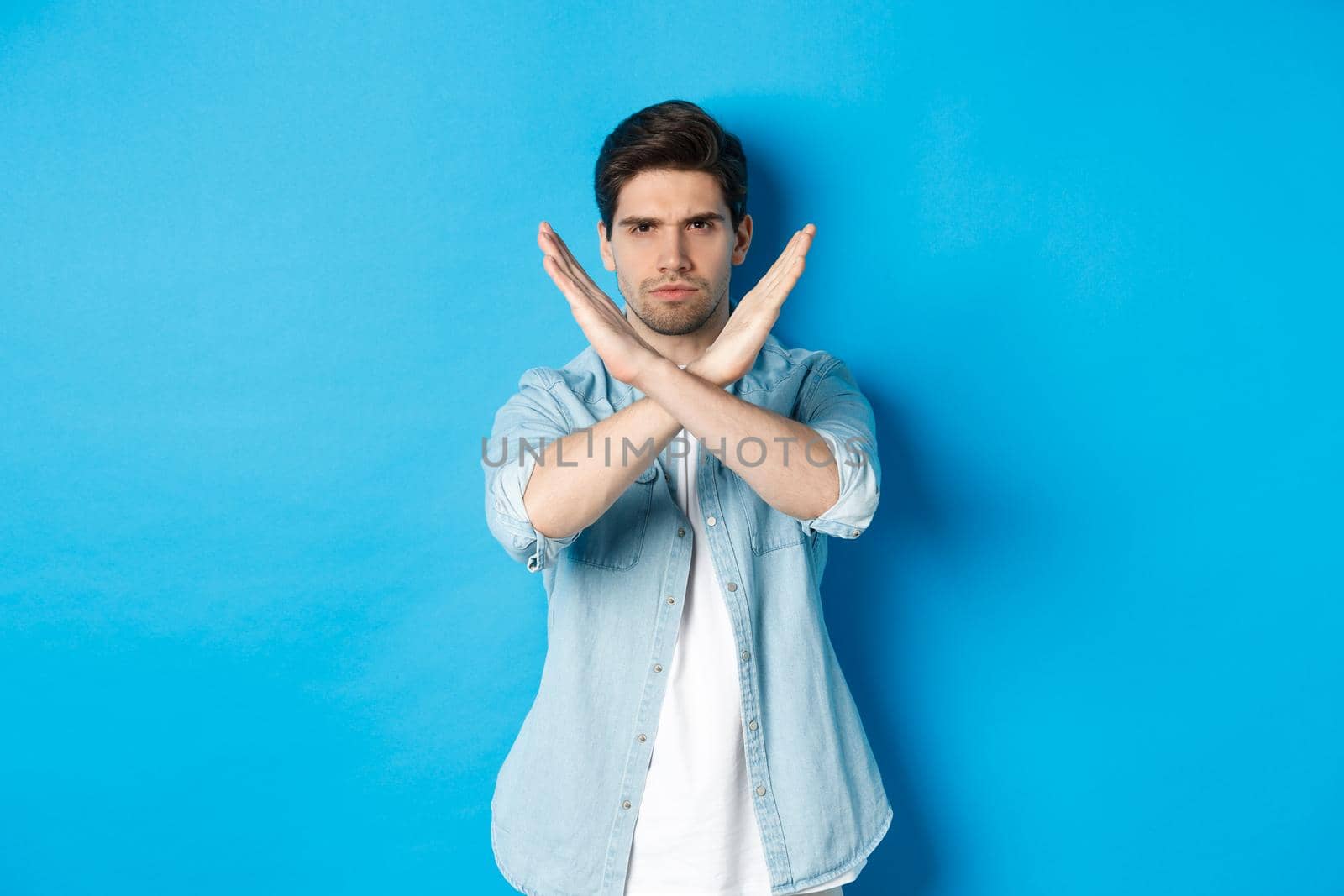 Displeased man frowning and saying no, making cross sign, tell to stop, standing against blue background.