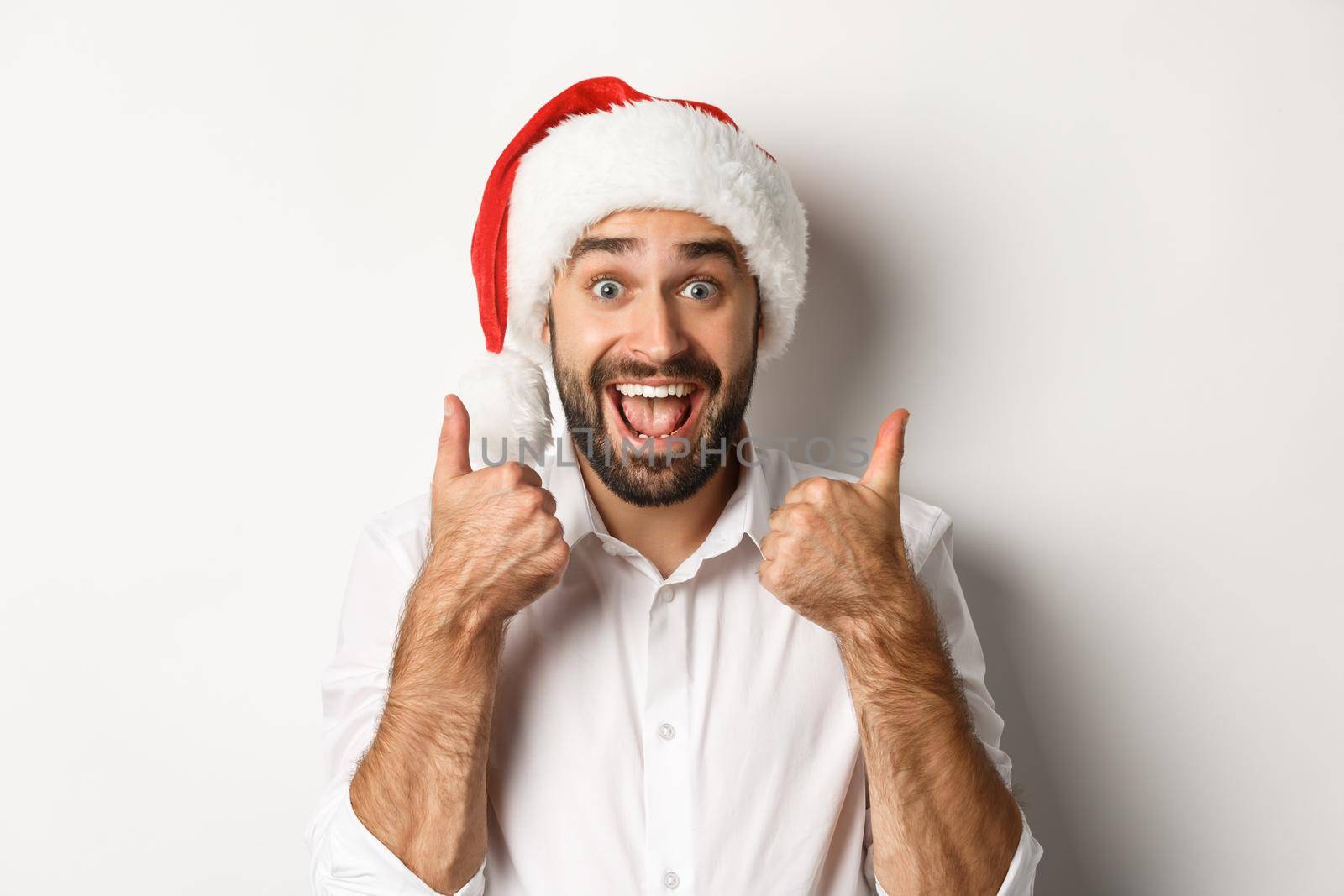 Party, winter holidays and celebration concept. Man enjoying christmas, wearing santa hat and showing thumb up with excited face, white background.