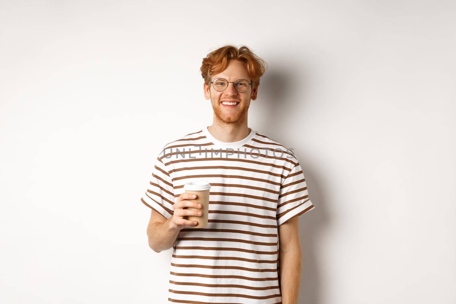 Handsome young man with beard and red messy hair, wearing glasses with striped t-shirt, drinking coffee from takeaway and smiling, white background.