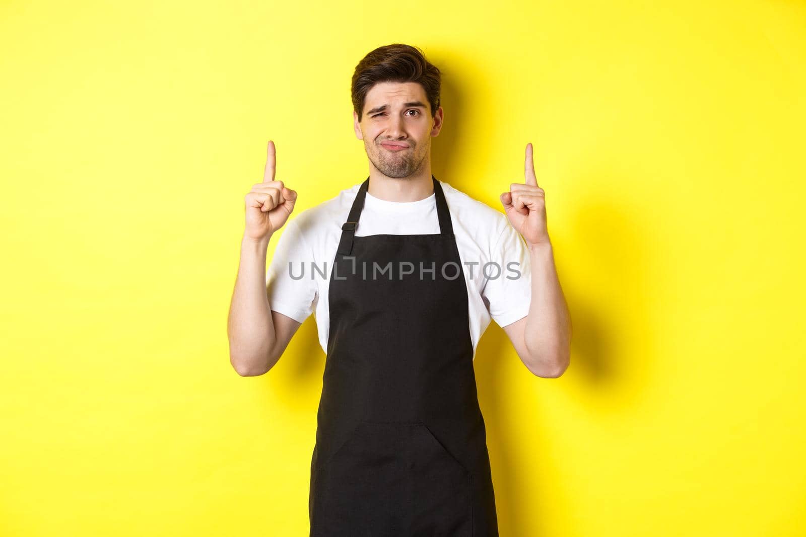 Displeased and skeptical waiter in black apron pointing fingers up, express dislike and indifference, standing over yellow background.