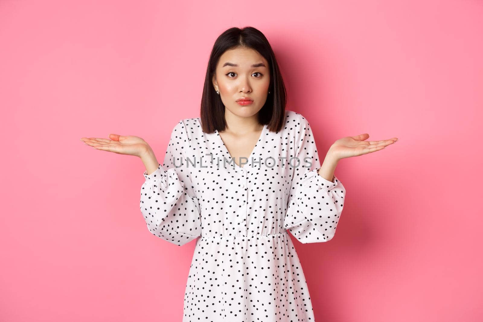 Indecisive cute asian woman shrugging, holding hands spread sideways on copy spaces, dont know, having doubts, standing over pink background.