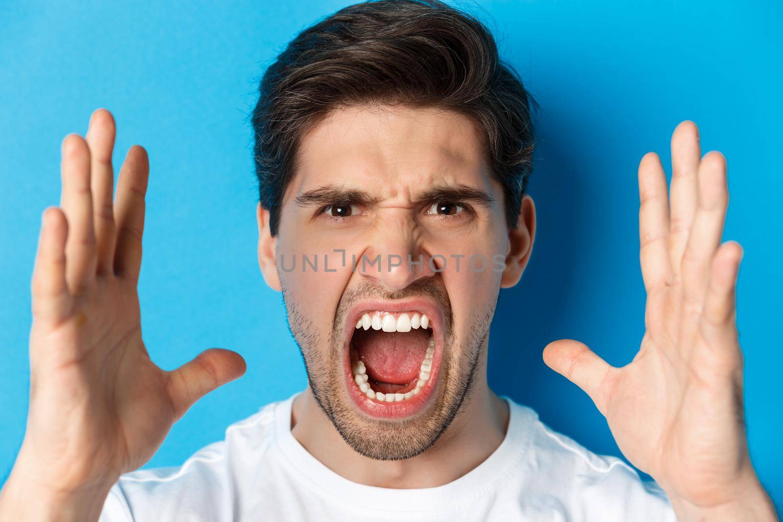 Headshot of man showing anger and frustration, yelling with outraged face, standing over blue background.