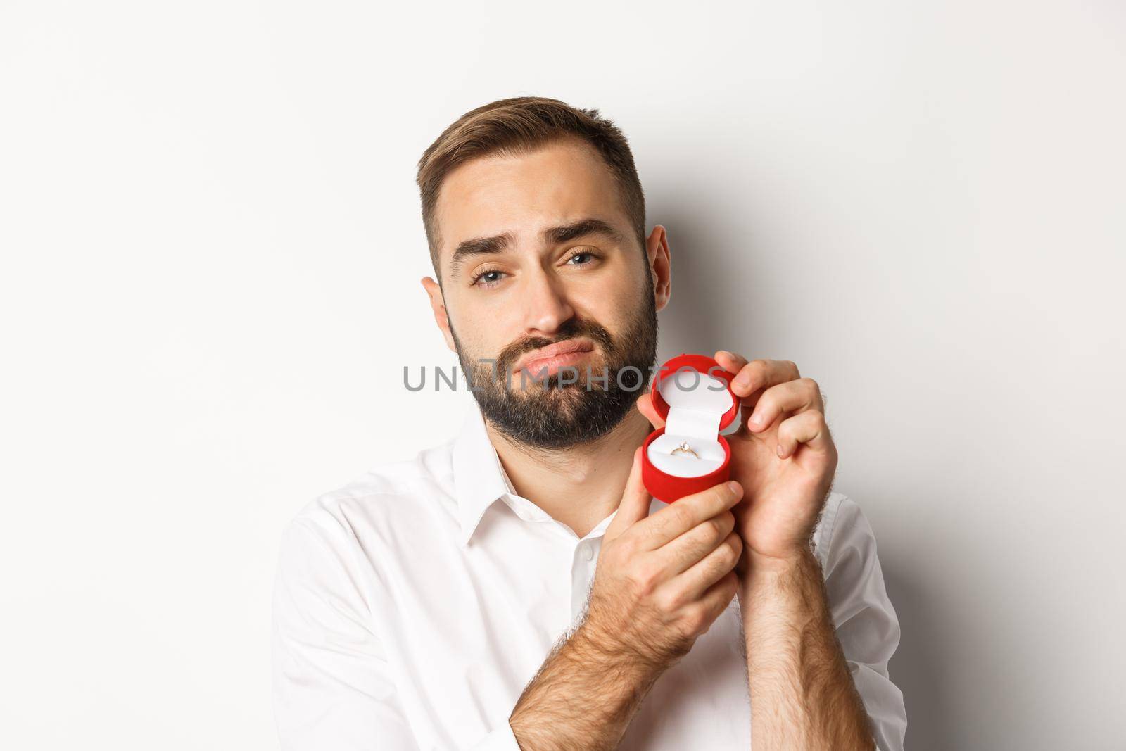 Close-up of hopeful man begging to marry him, looking sad and showing wedding ring, making a proposal, white background.