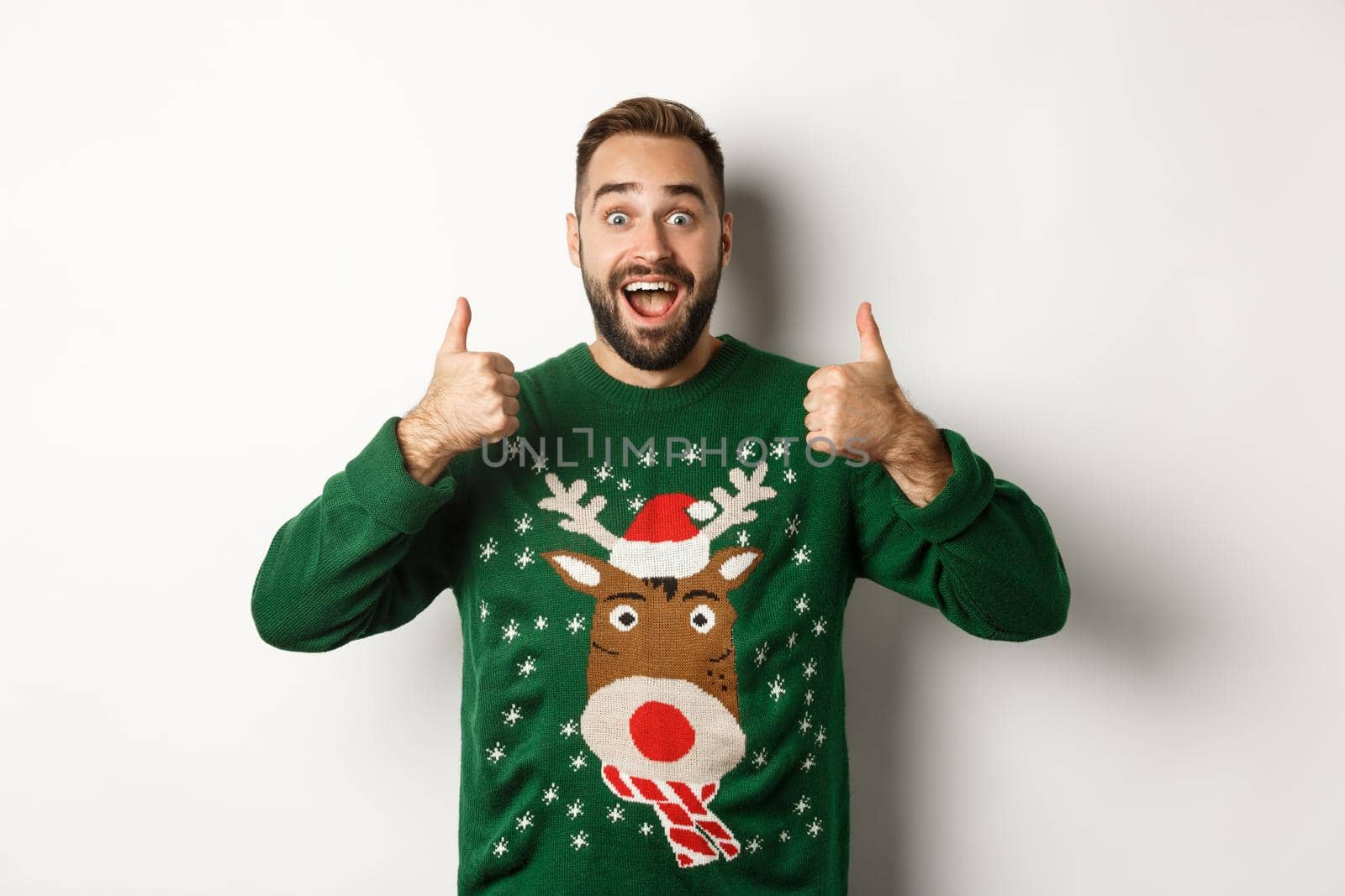 Christmas, holidays and celebration. Excited bearded guy in green sweater showing thumbs up, like something amazing, standing over white background.